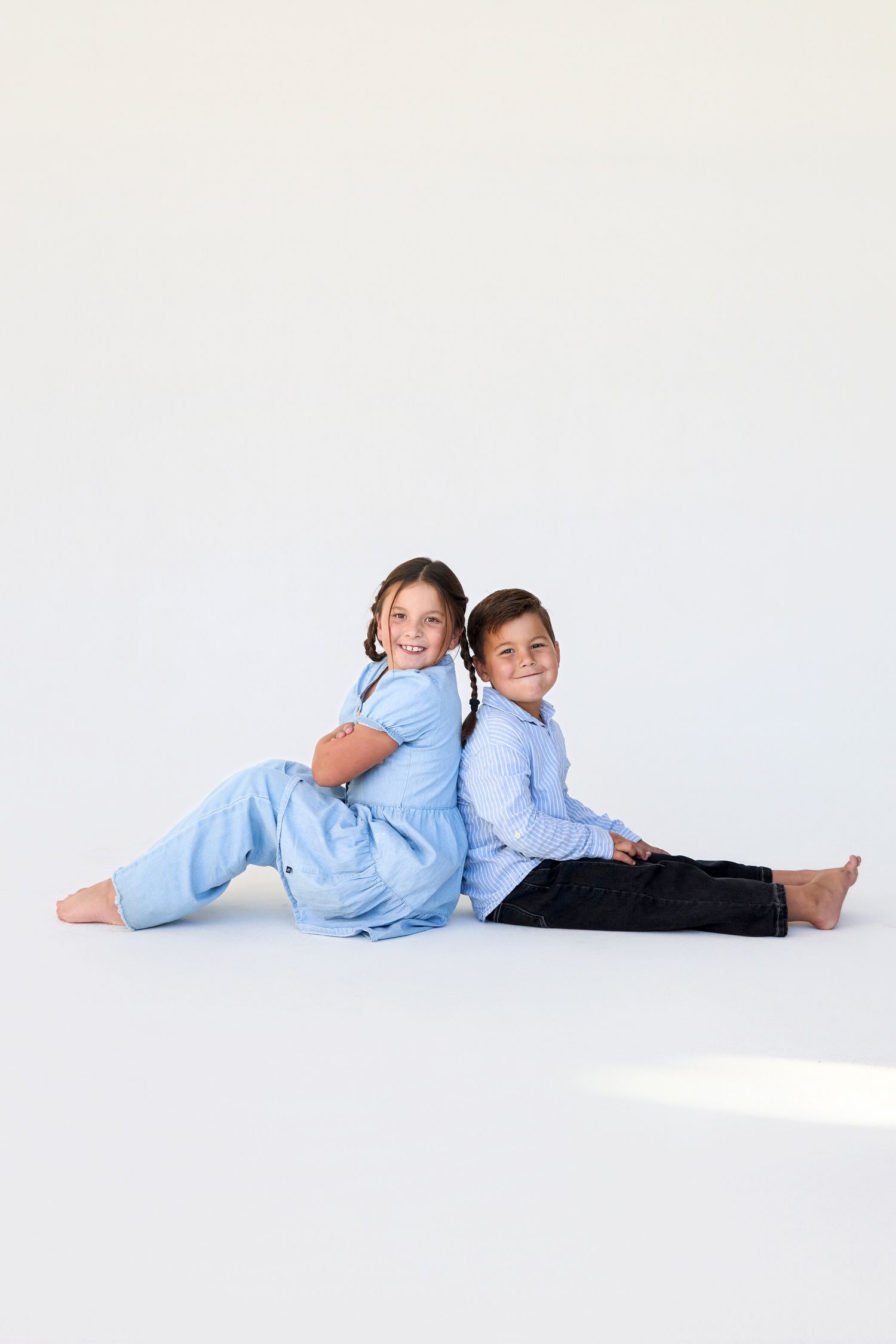 Two children in blue outfits sit back-to-back on the floor, smiling at the camera against a white backdrop.