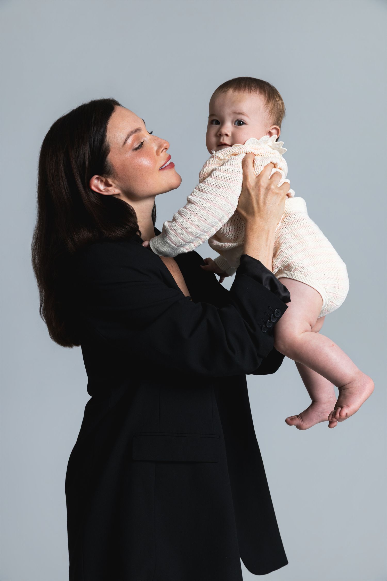 A woman in a black blazer joyfully holds a smiling baby in a white outfit against a gray background.