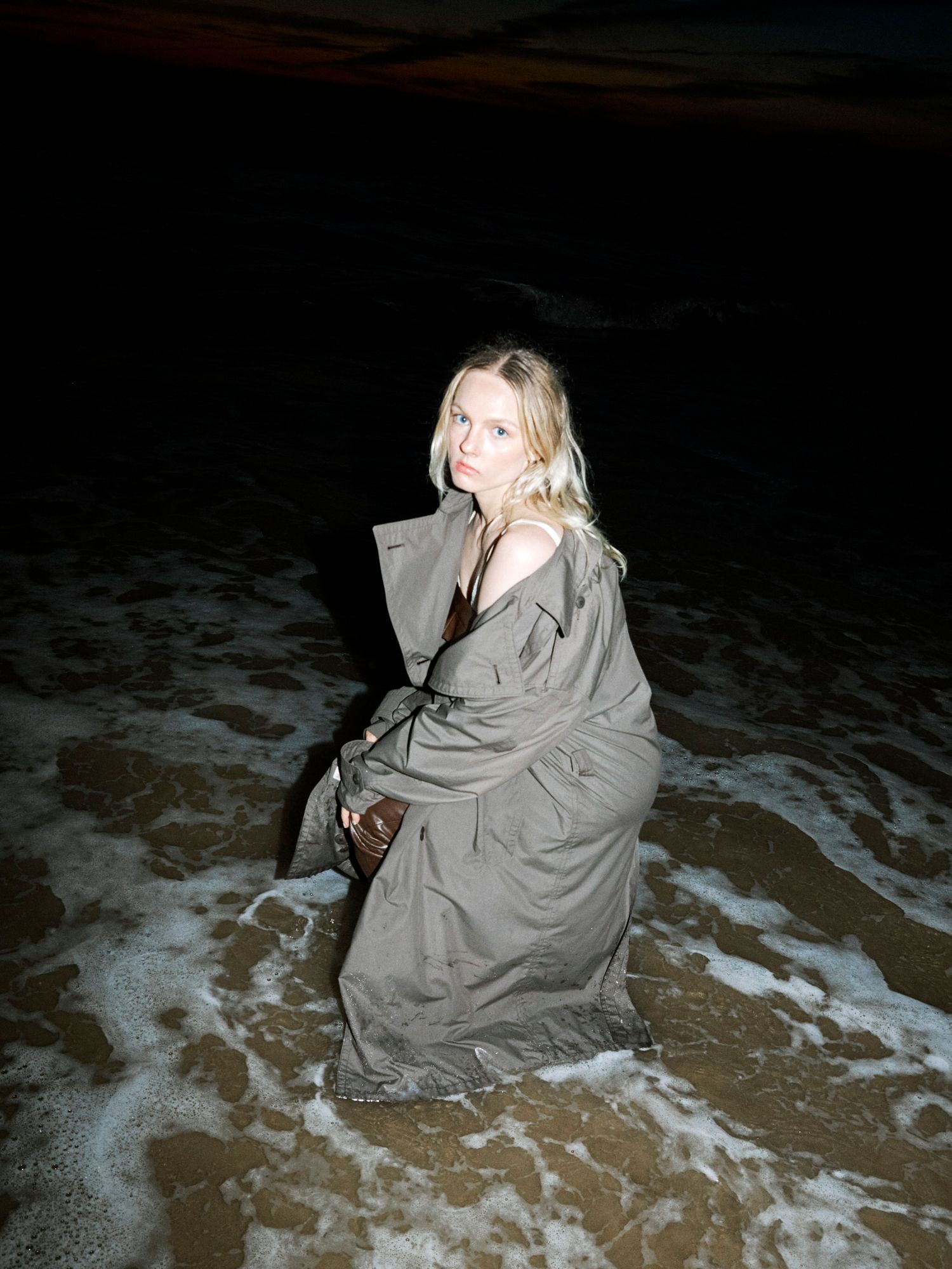 Blonde woman wrapped in olive trench coat crouching on sandy beach with waves at night.