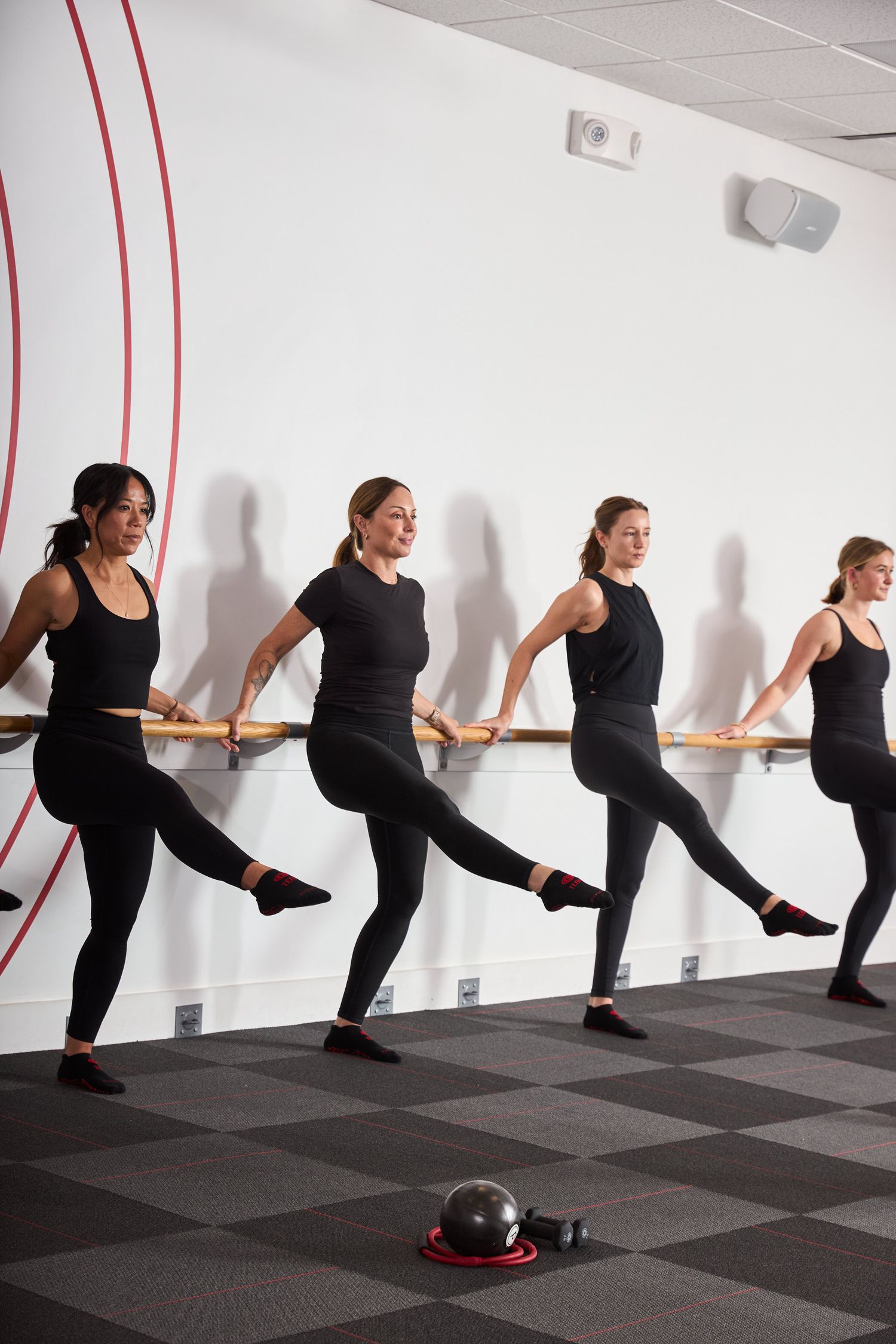 Four women in black workout attire practicing ballet barre exercises in a white fitness studio.