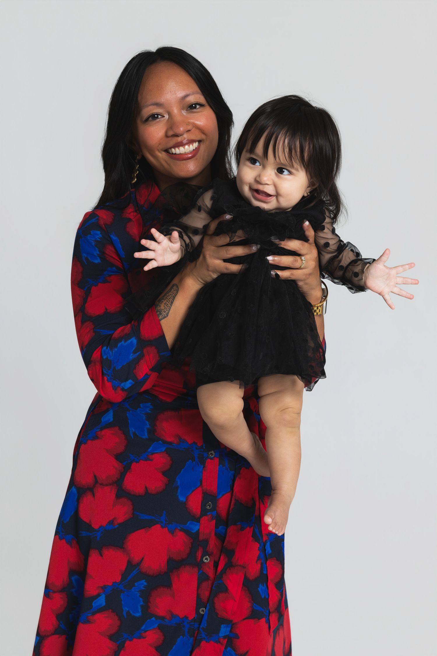 Smiling woman in floral dress holding a baby girl in black dress against white background.