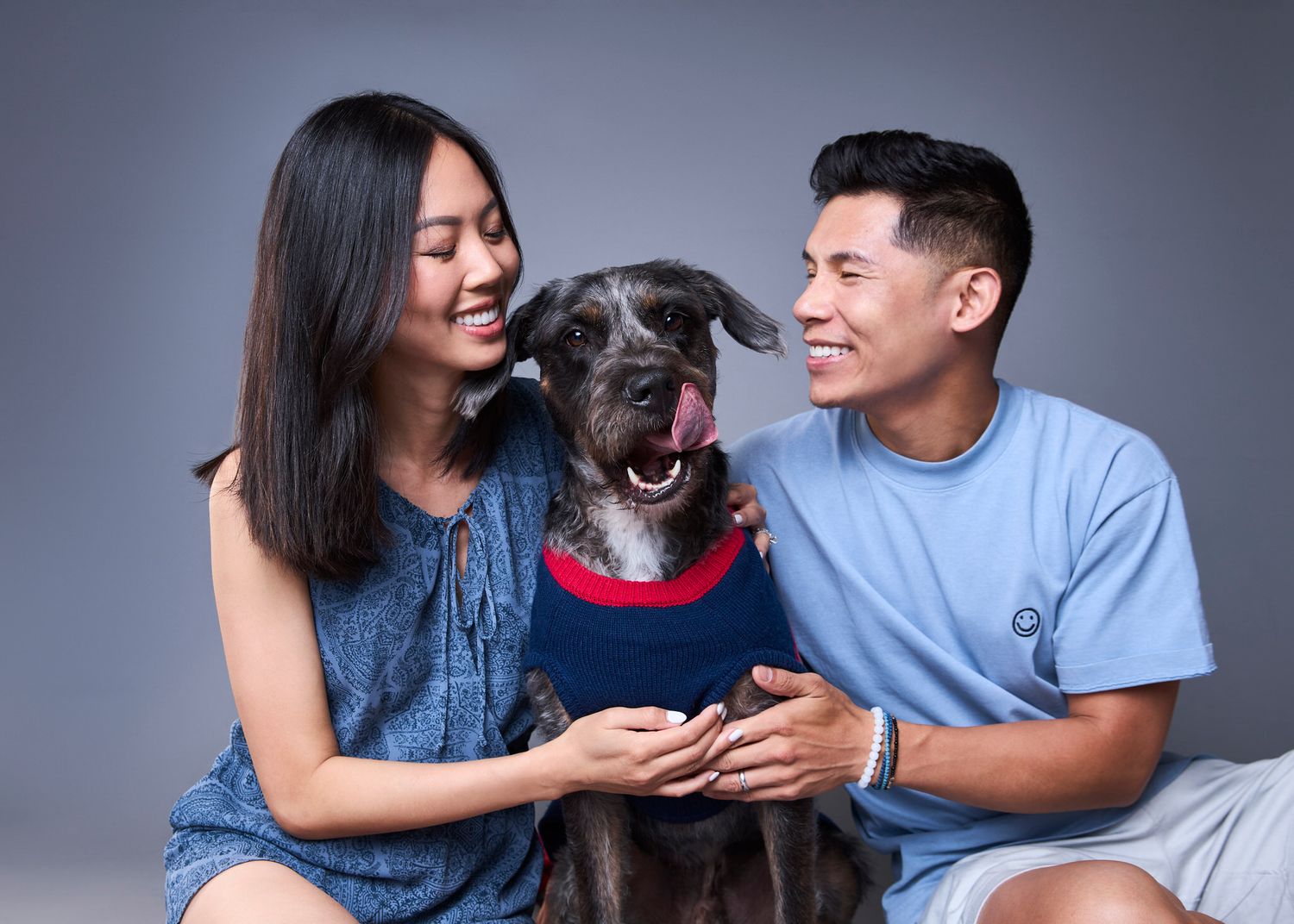 A smiling couple poses with their happy black dog wearing a red and blue shirt against a gray background.