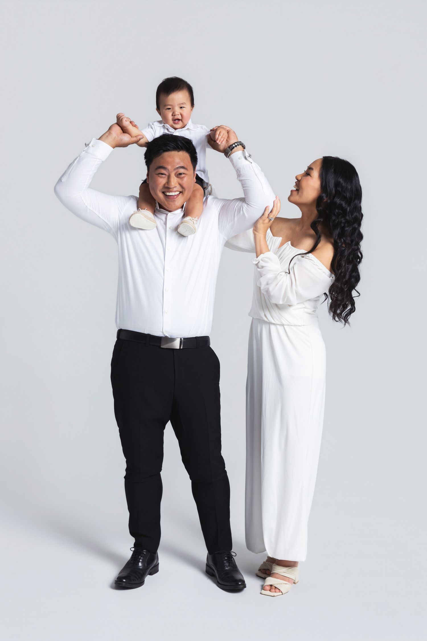 Happy Asian family portrait with father carrying toddler on shoulders, mother in white dress, studio photo.