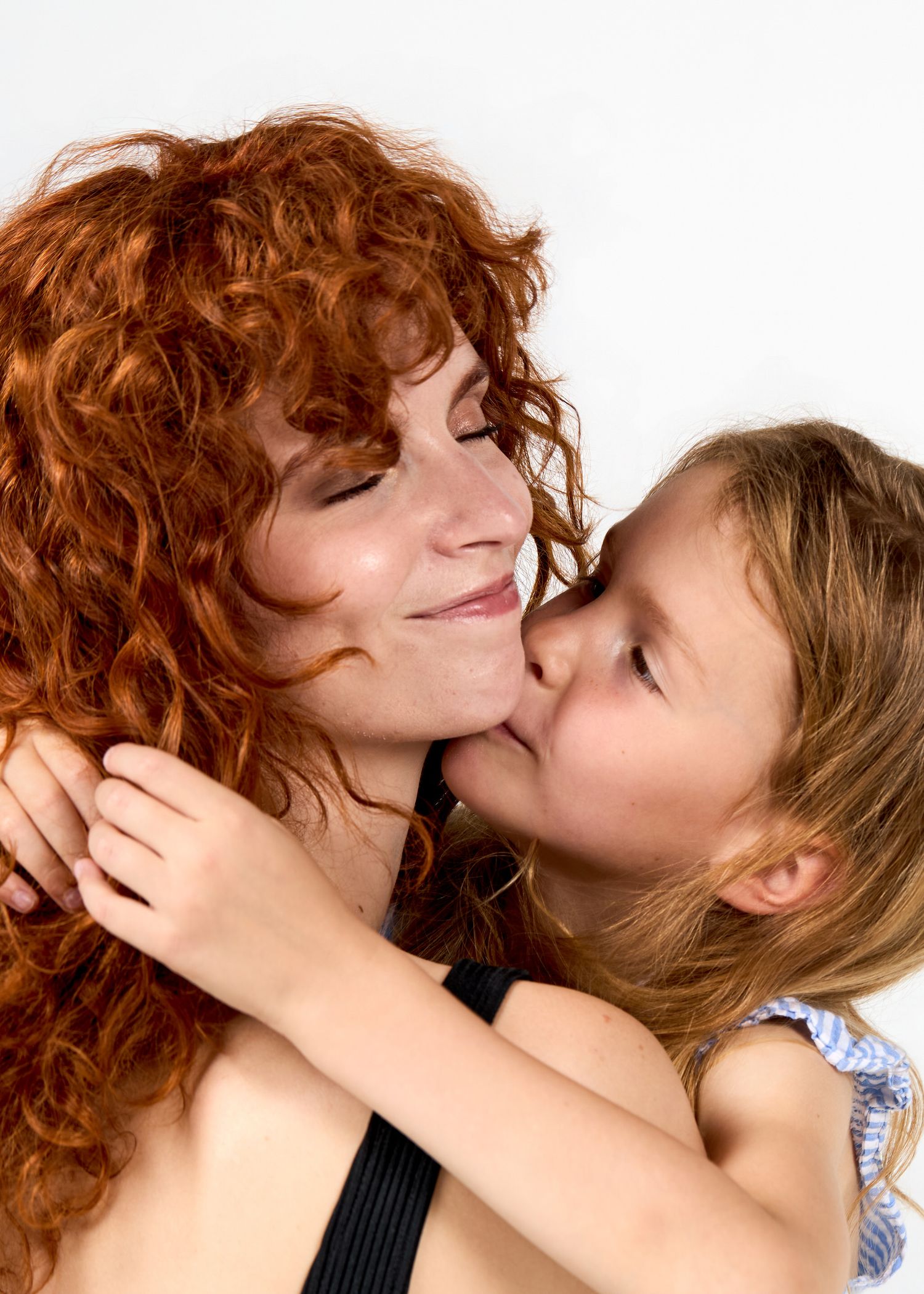A young girl gives a tender kiss on the cheek to her curly redhead mother in a studio portrait.