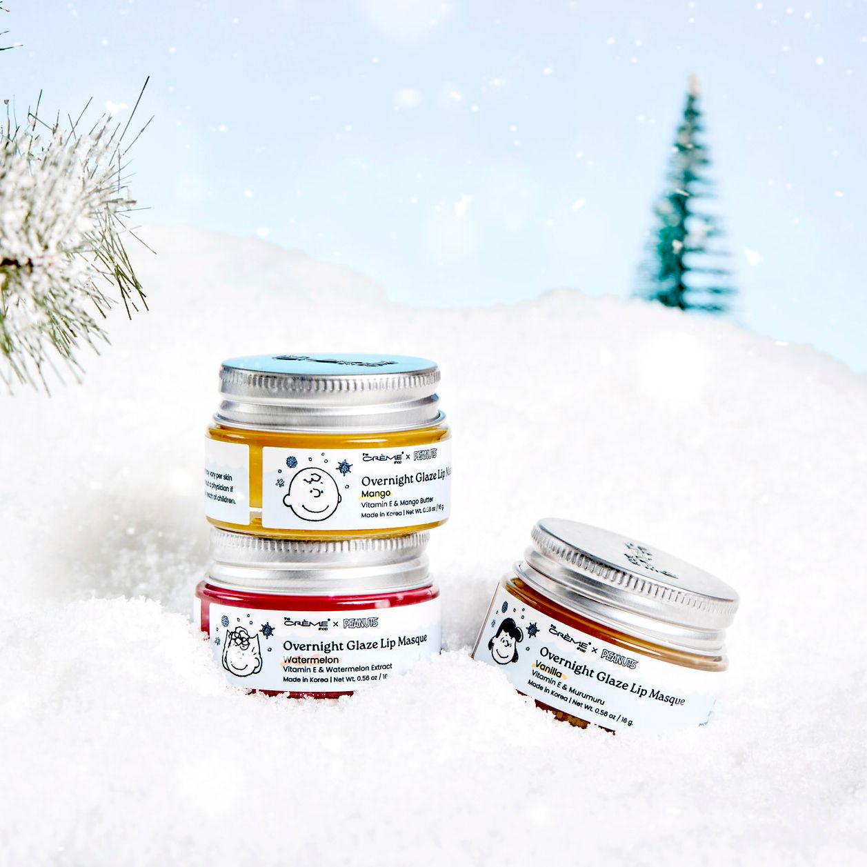 Three Overnight Glaze Lip Masque jars in mango and other scents displayed in a snowy winter scene with pine trees.