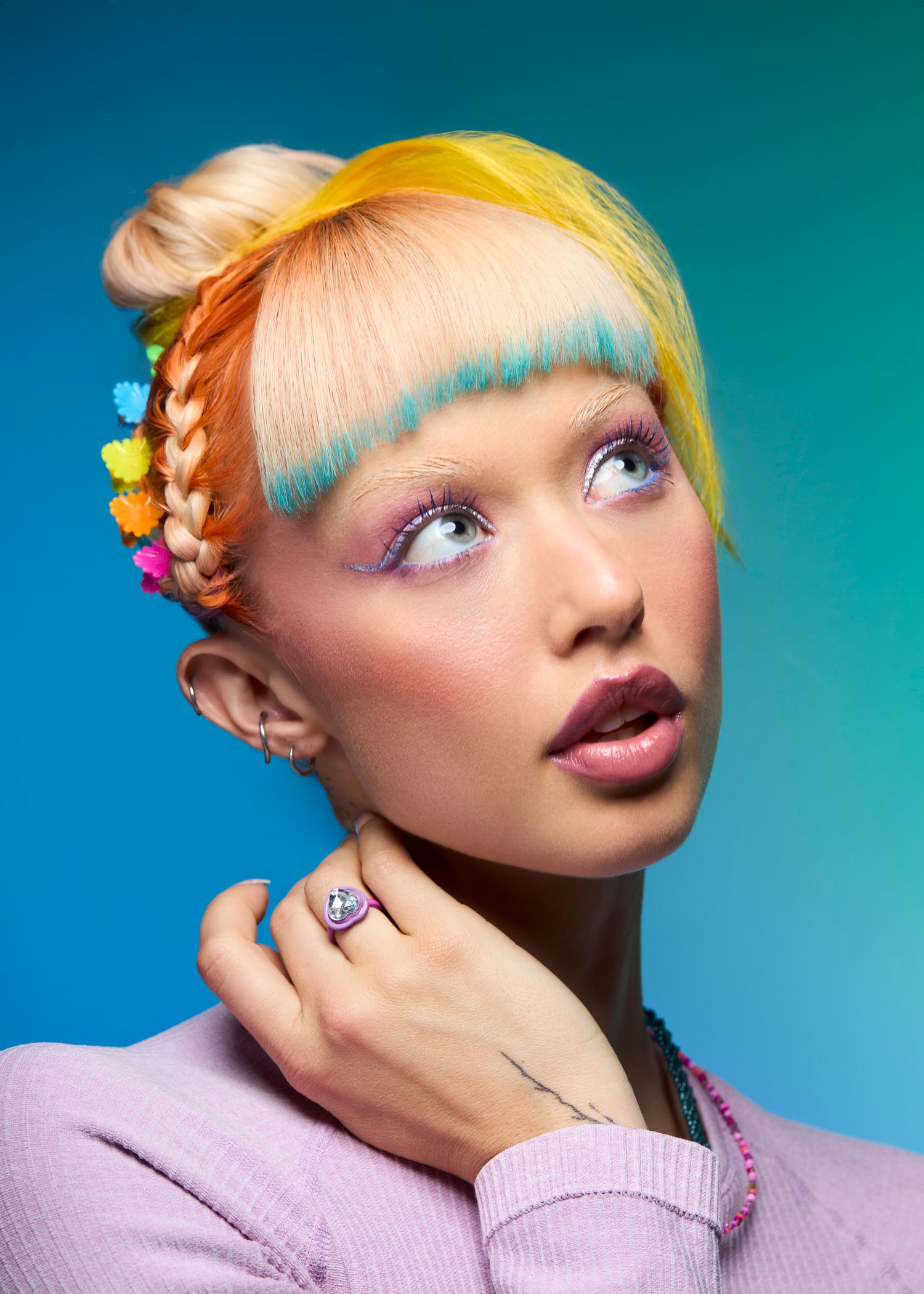 Model with multicolored hair featuring teal bangs, orange braid, and yellow top knot adorned with colorful flower clips.
