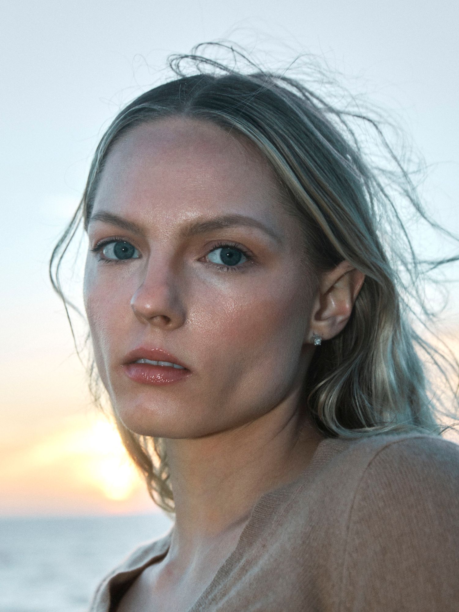 Young woman with blonde hair and blue eyes poses near ocean at sunset in a candid outdoor portrait.