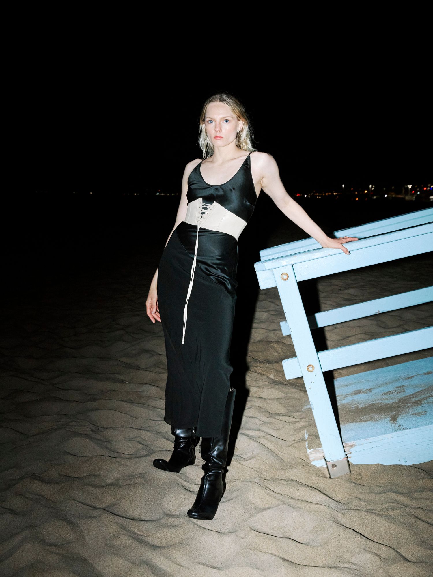 Woman in black satin crop top and midi skirt with corset detail standing on beach at night.