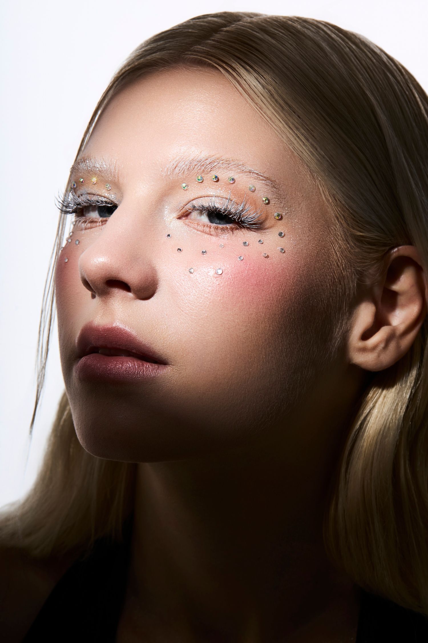 Close-up of a model with rhinestone embellishments, rosy blush, and dramatic white eye makeup in an artistic beauty look.
