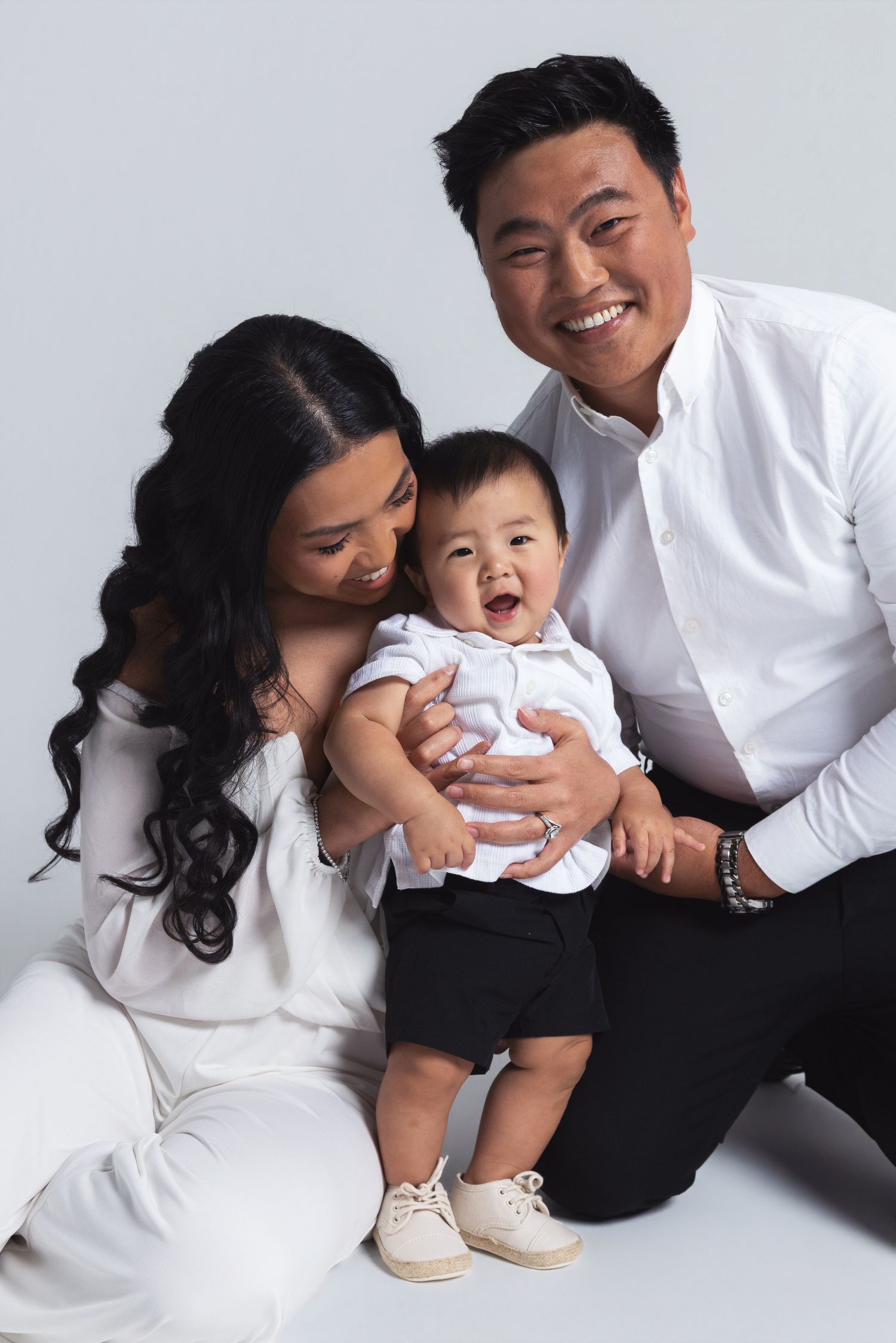 Happy Asian family portrait with smiling parents and adorable baby boy dressed in white and black on light background.
