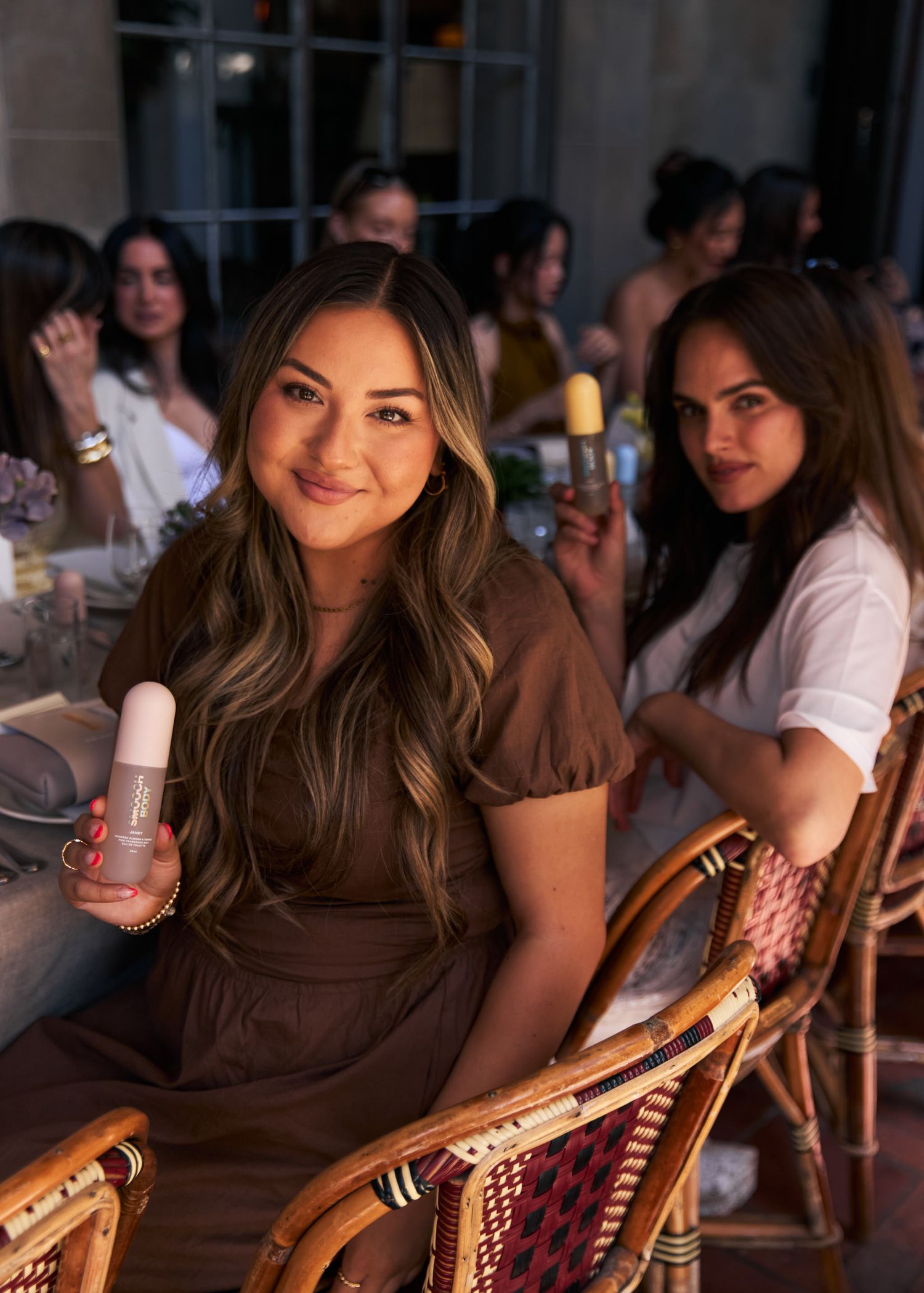Two women at a dinner party holding skincare products, smiling at camera in warm evening light.