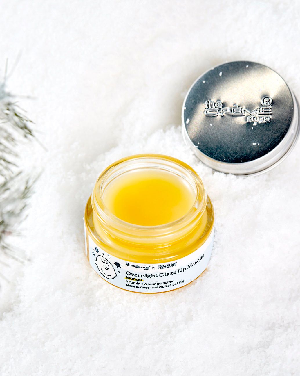 Open jar of Overnight Glaze Lip Mask with golden balm, silver lid, on white snow with pine branches.