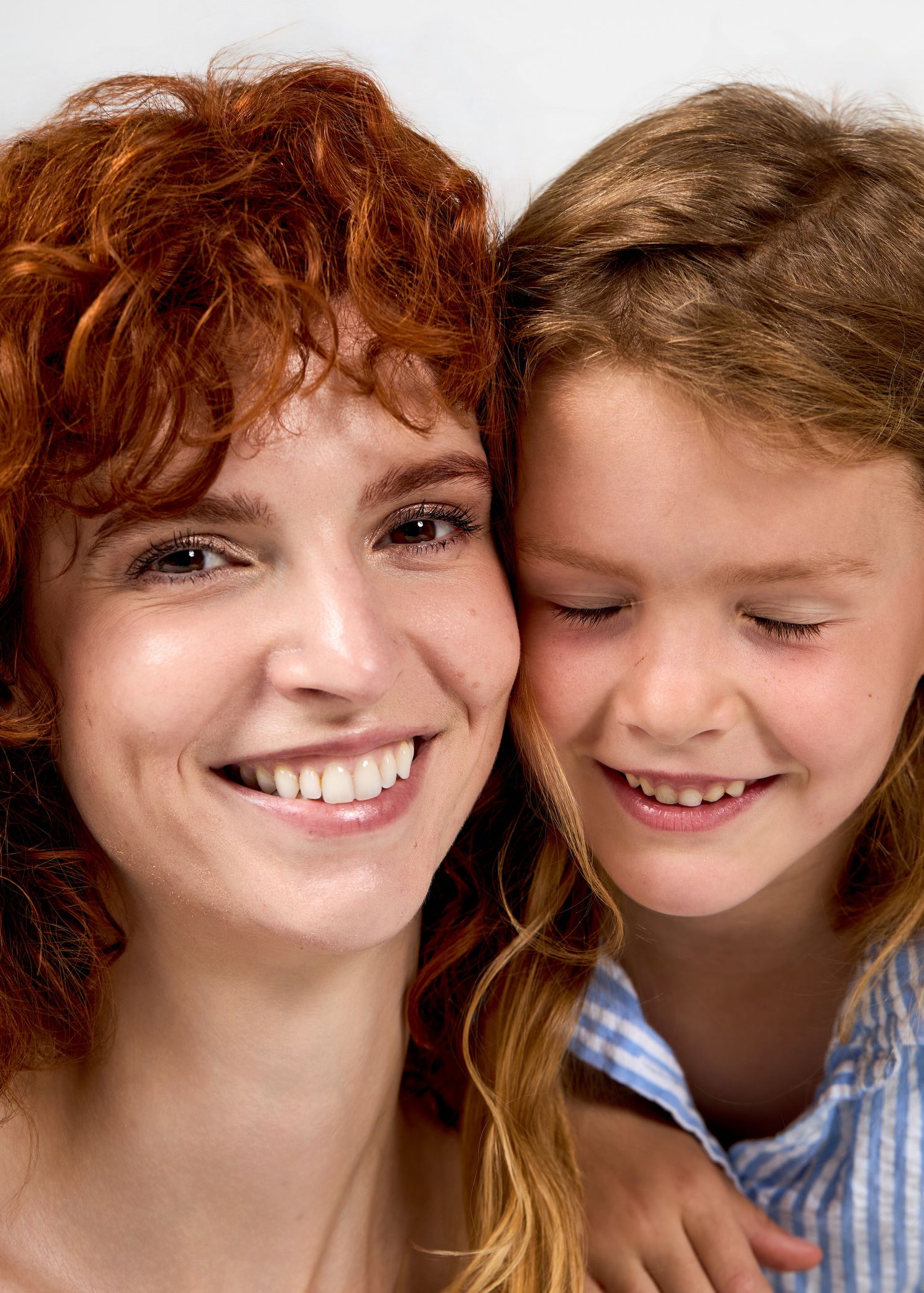 A smiling redhead woman and a young girl with light brown hair share a joyful close-up moment together.
