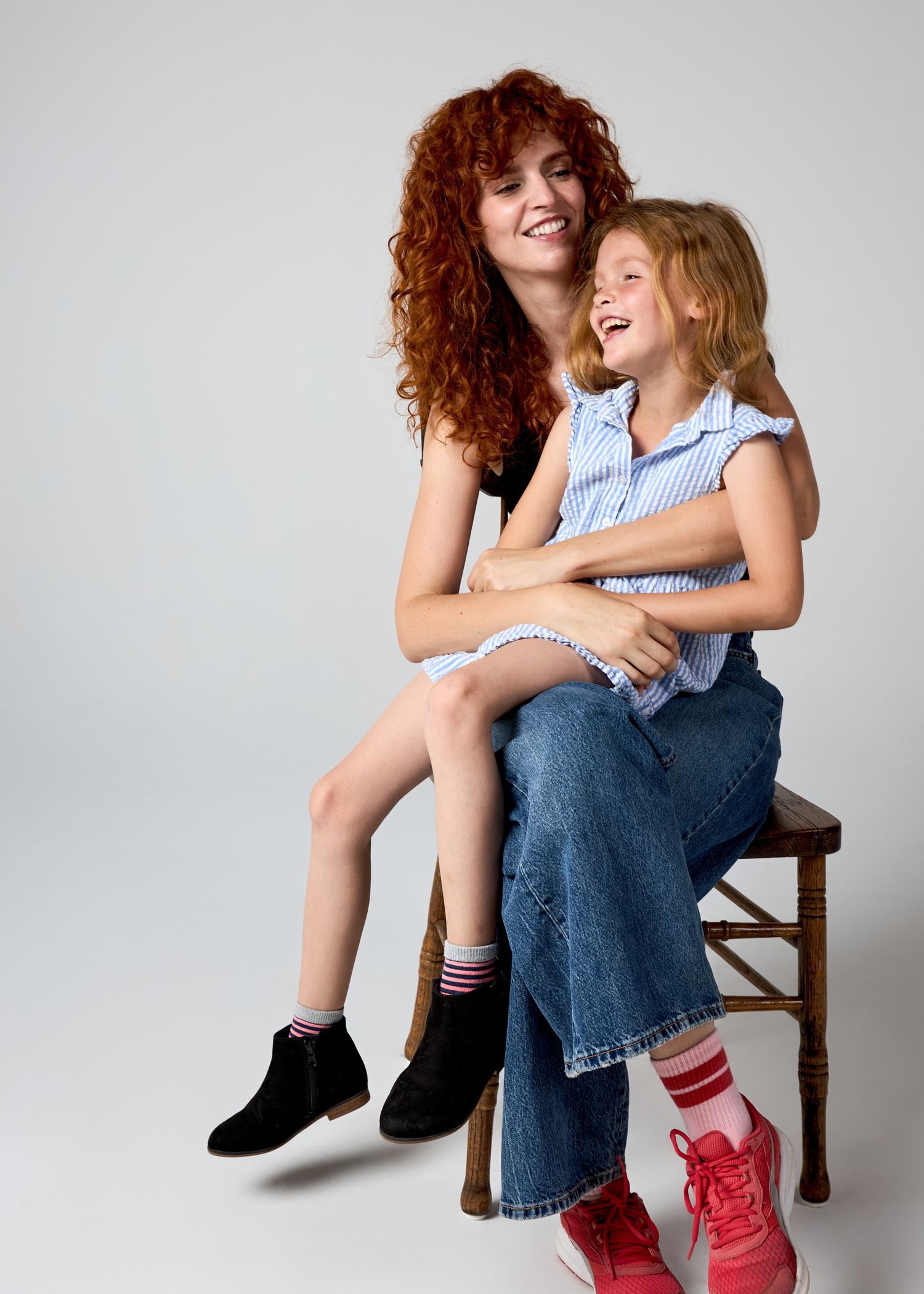 A curly redhead mother hugs her laughing daughter on a wooden stool in a studio portrait.