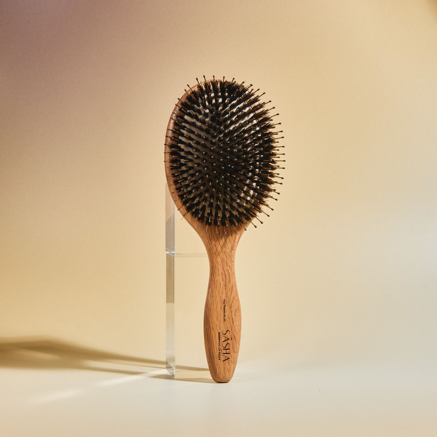 Oval wooden paddle hair brush with boar bristles displayed upright on a warm beige background.