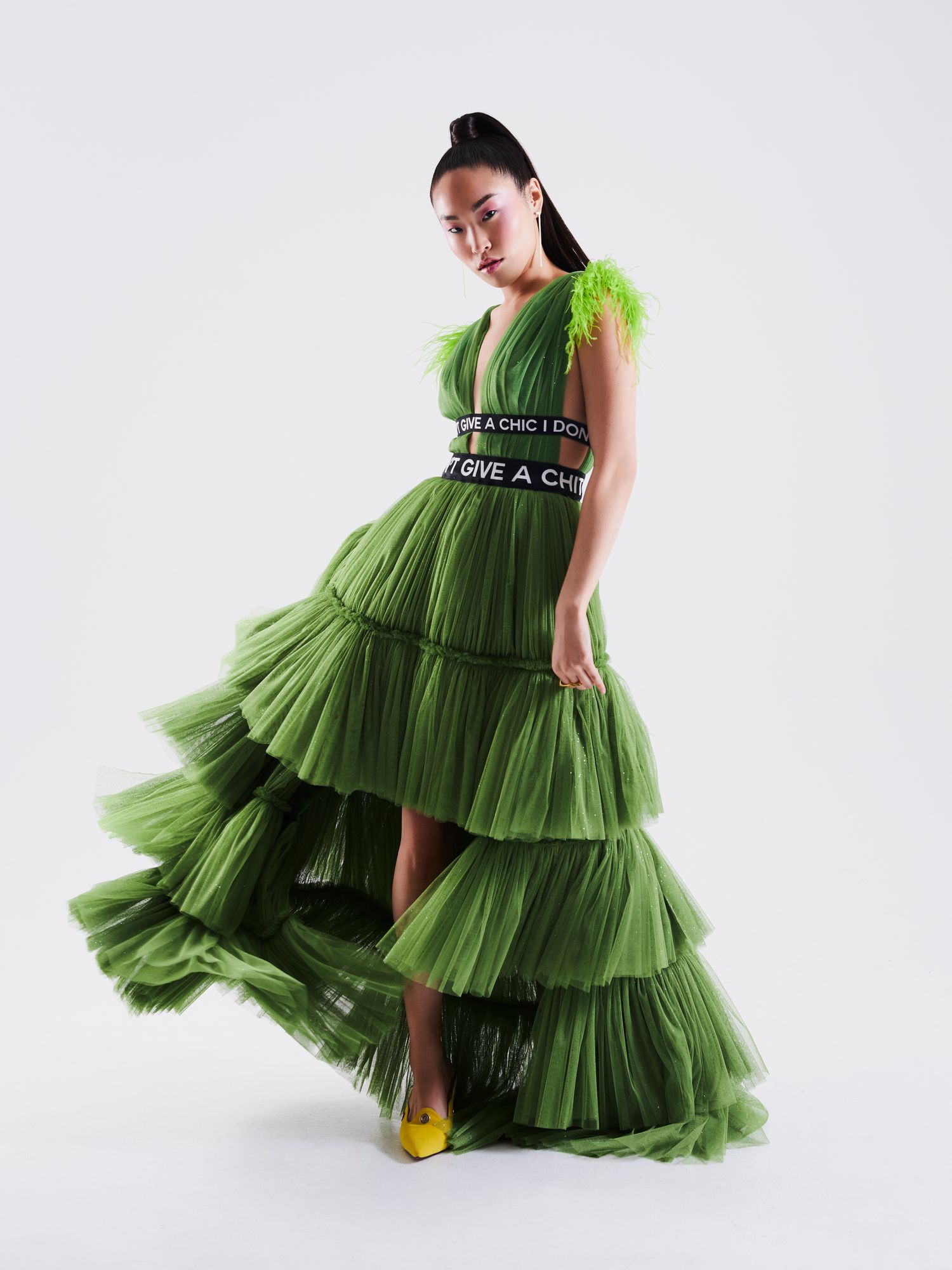 Model wearing a green tiered tulle gown with feather accents and a logo waistband, paired with yellow heels.