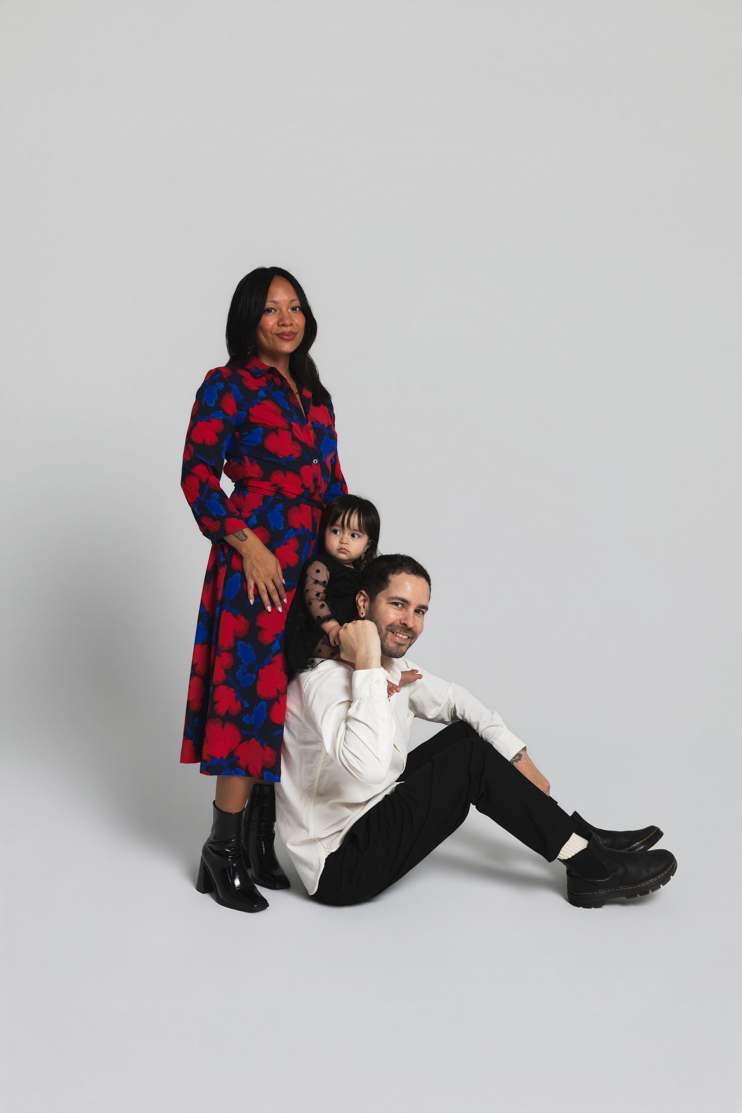 A family of three poses in a studio, woman in floral dress, man seated, child between them.