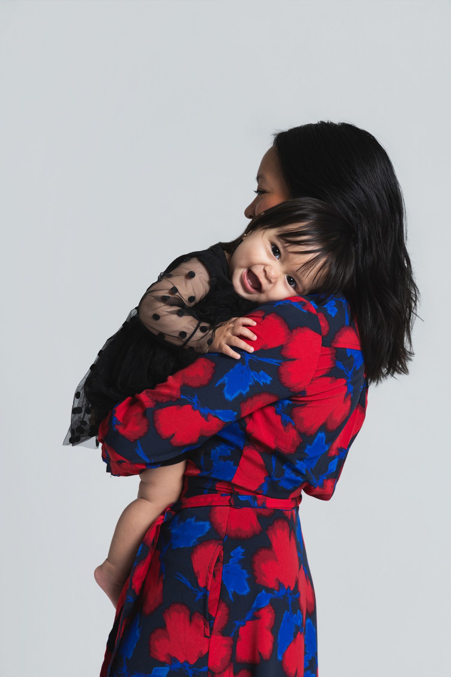 Woman in floral red and blue dress holding a smiling toddler in a black outfit against a grey background.