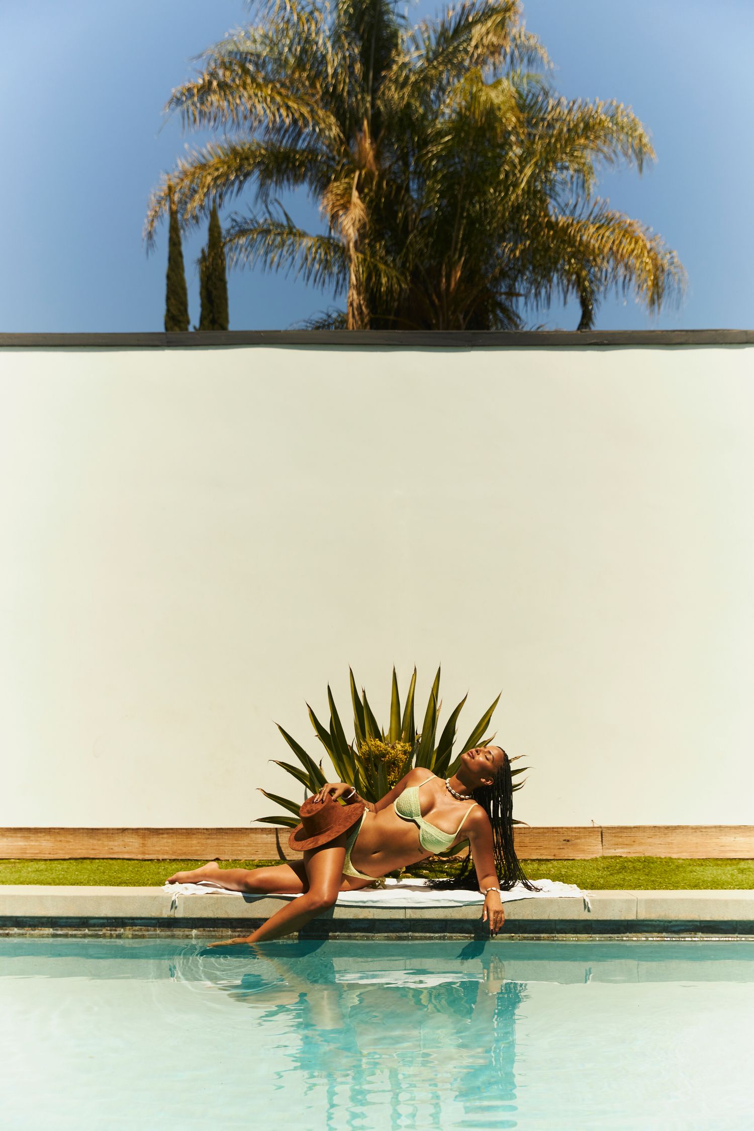 Woman in green bikini sunbathing by a pool with palm trees and white wall backdrop on a sunny day.