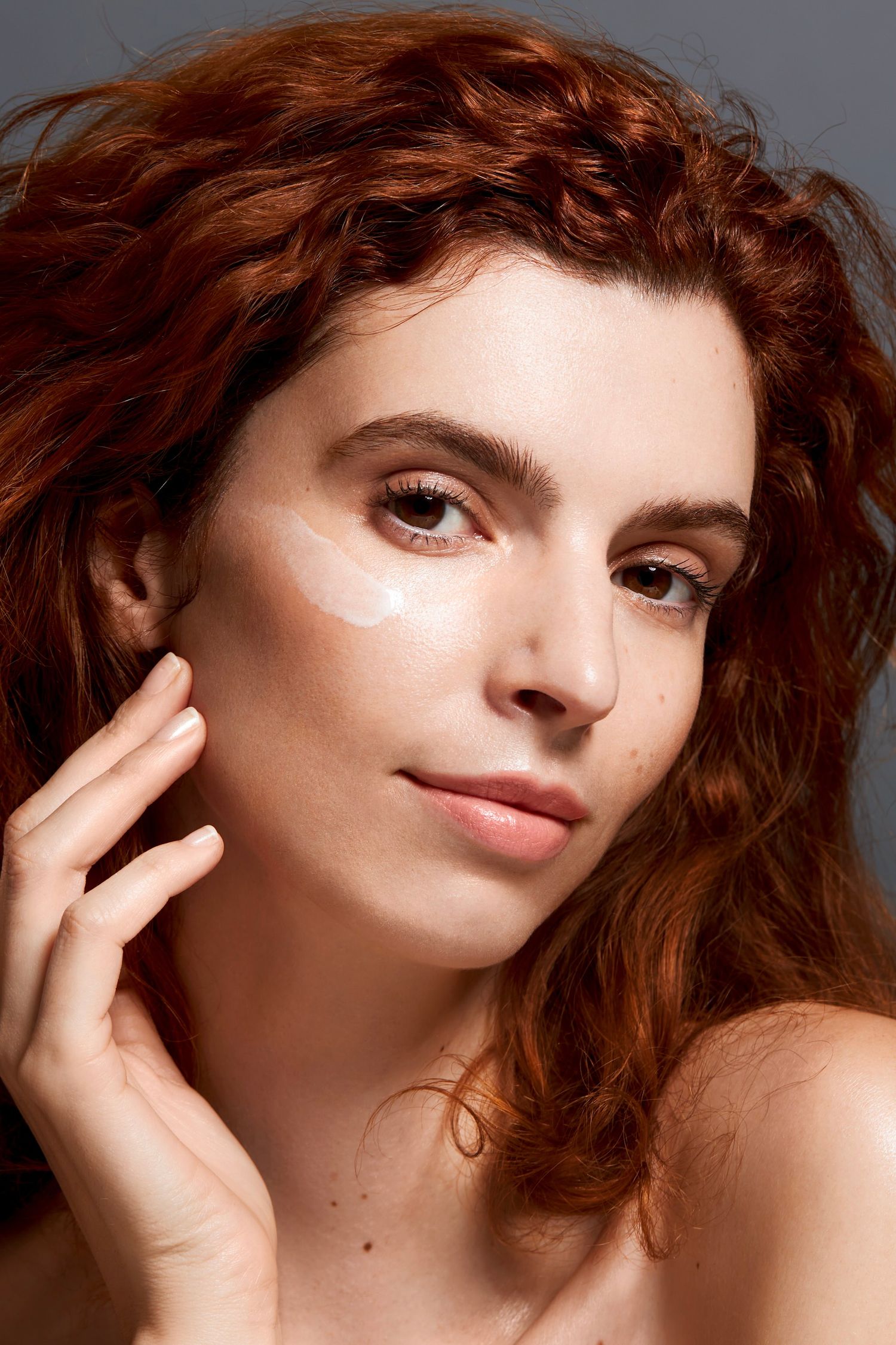 A redhead woman with curly hair applying white face cream to her cheek in a beauty skincare routine.