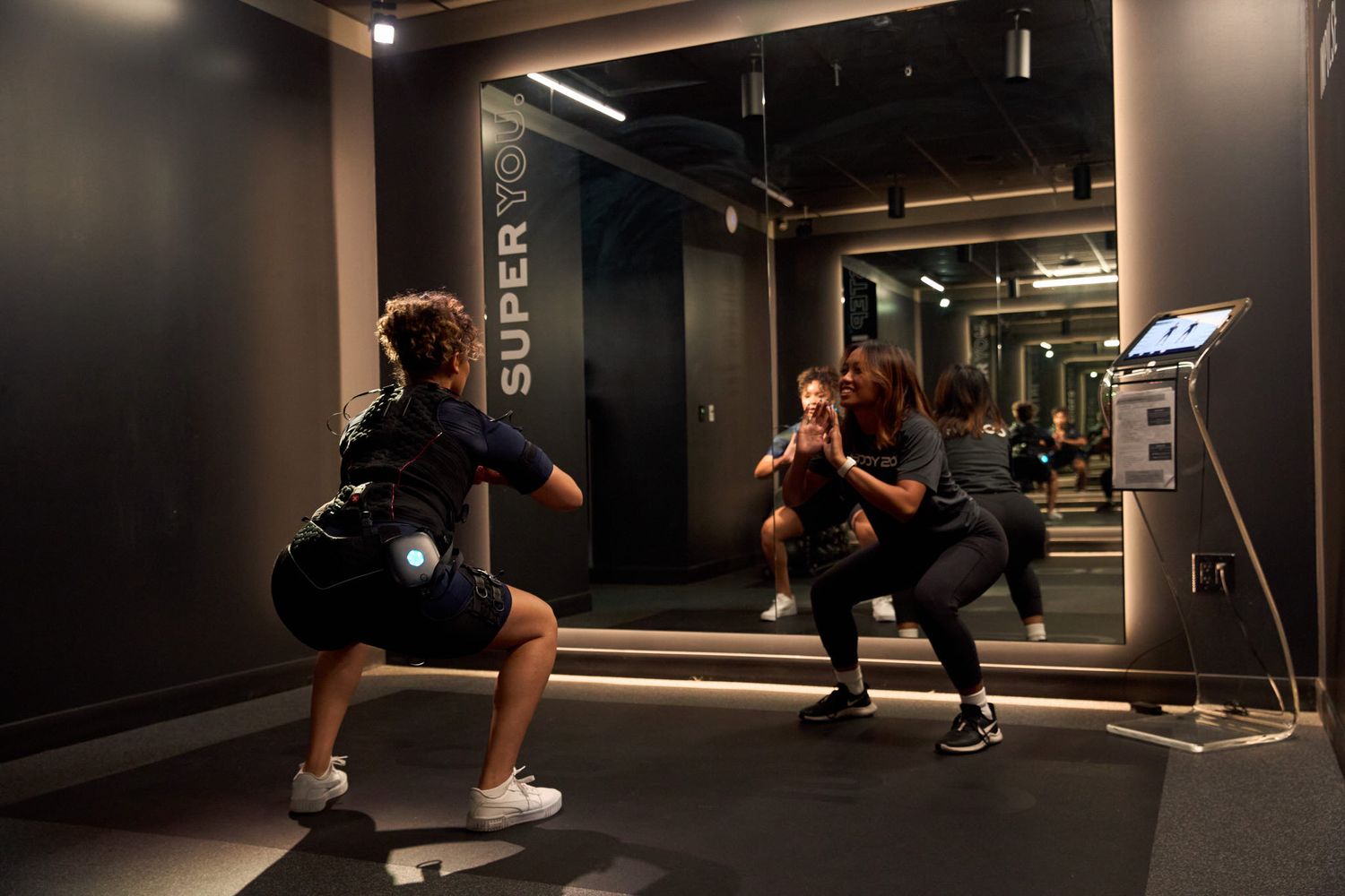 Two people performing squat exercises with a trainer in a modern gym with mirrored walls and EMS training suits.