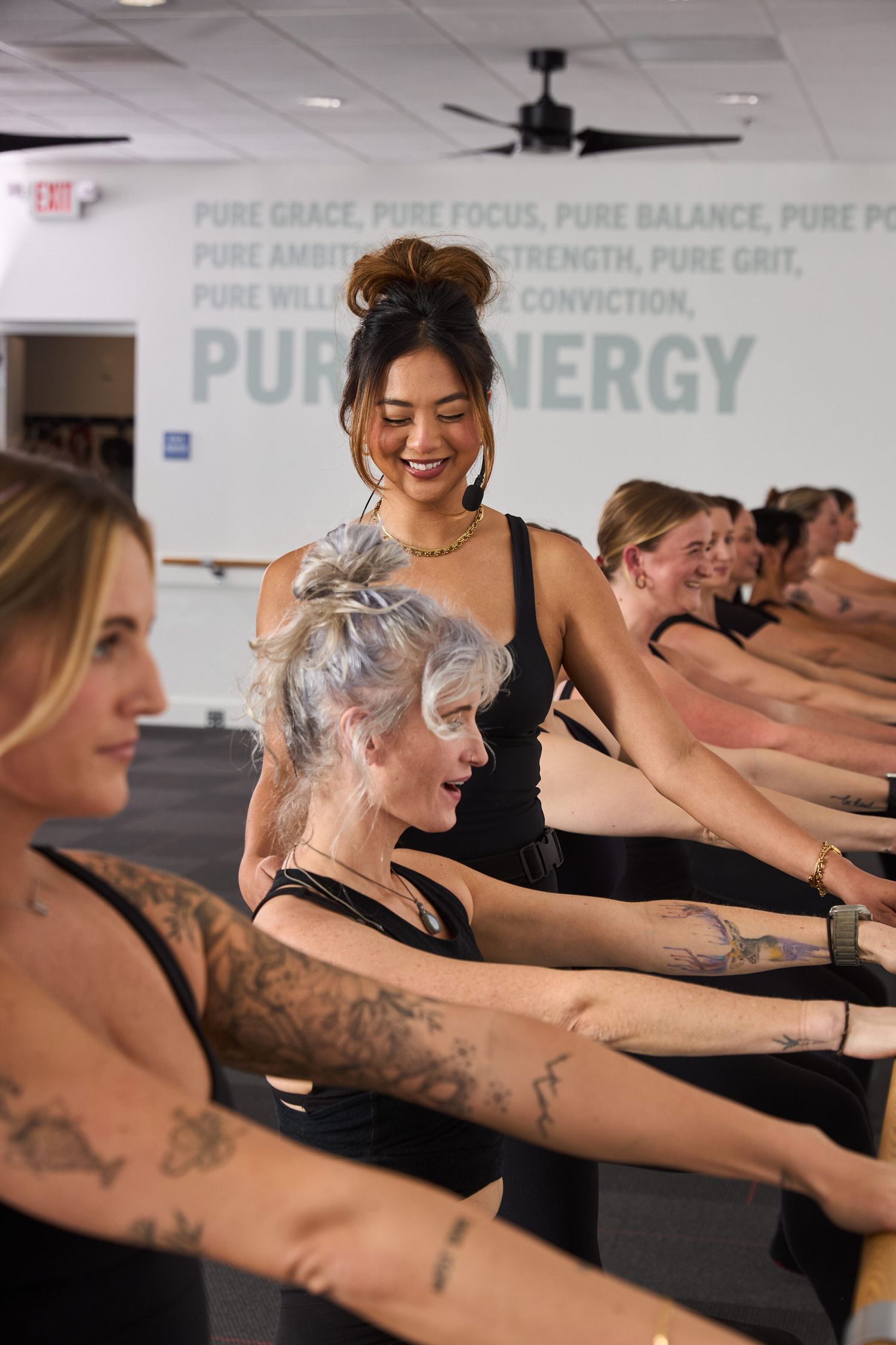 Women in black activewear practice barre exercises in a fitness studio with Pure Energy motivational wall text.