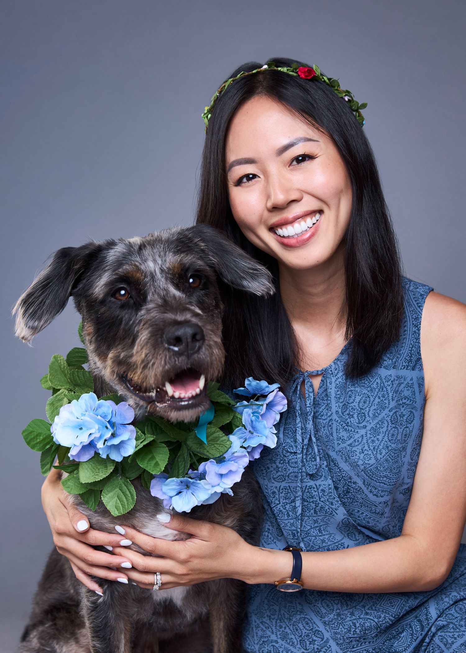 Smiling woman wearing a floral crown poses with a fluffy dark dog adorned with blue hydrangea collar in studio.