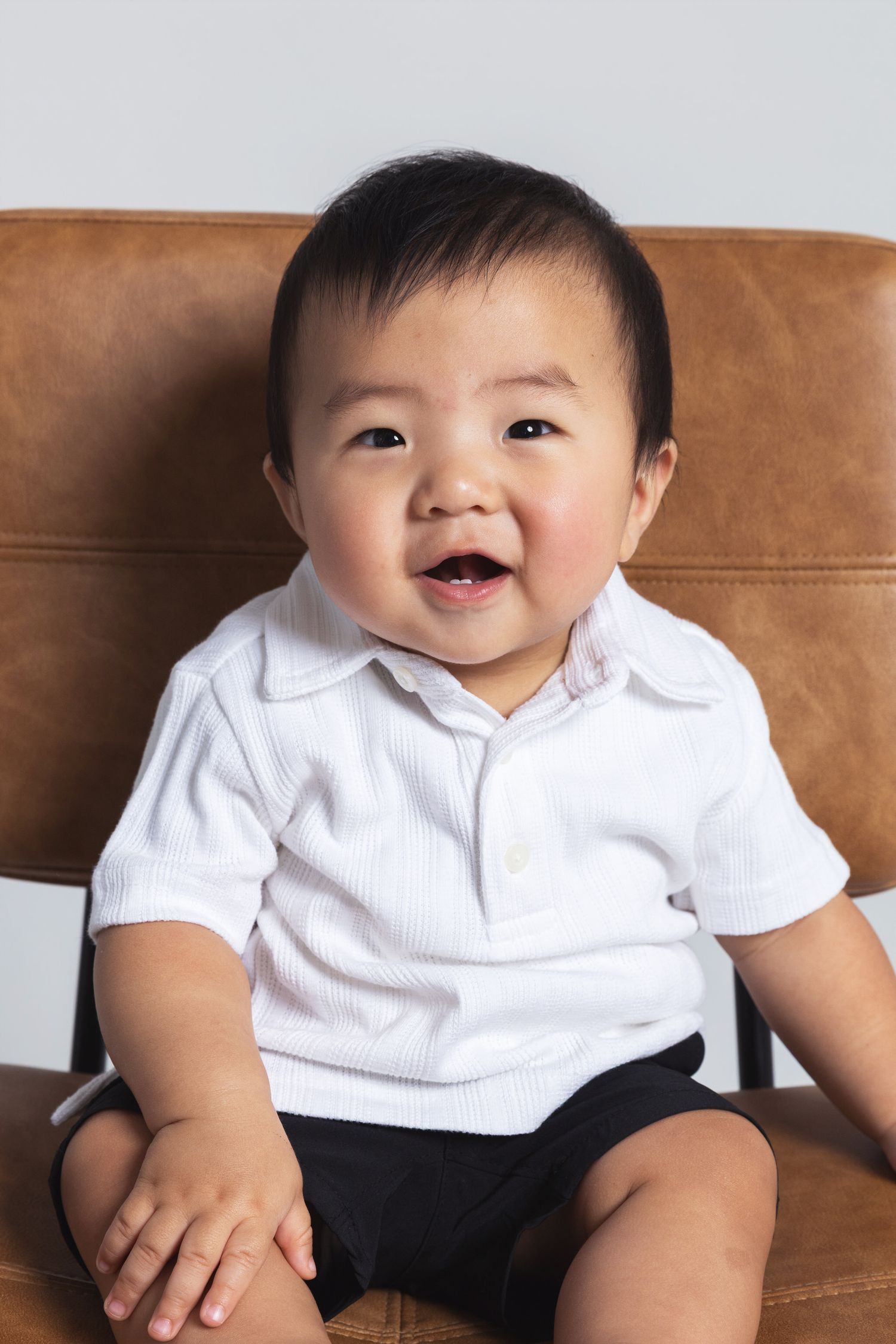 Smiling Asian baby in white polo shirt and black shorts sitting on brown leather chair.
