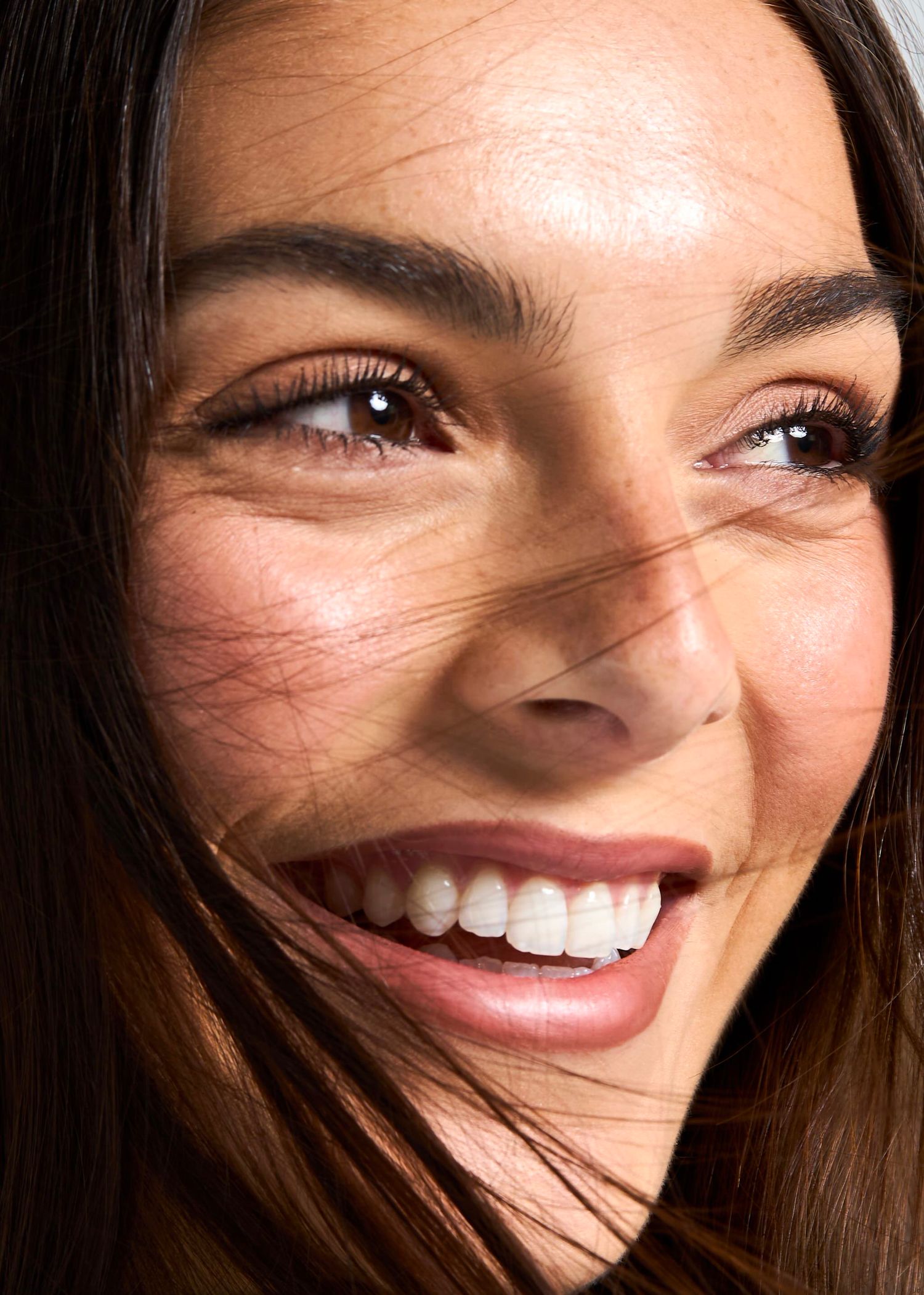 Close-up of a smiling woman with brown eyes, glowing skin, and dark hair blowing across her face.
