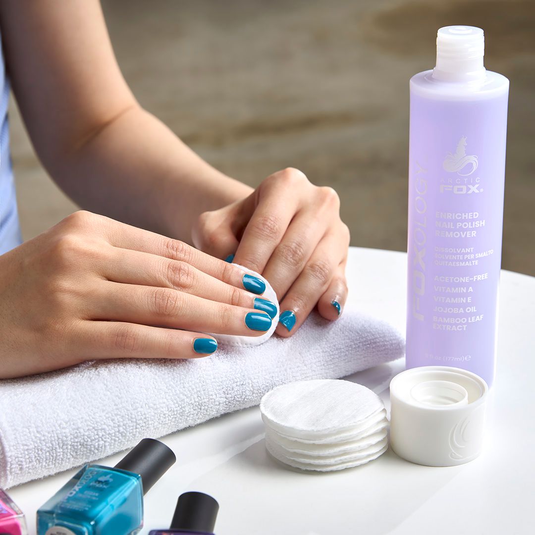 Woman removing teal nail polish with OXX acetone remover, cotton pads, and towel on white table.