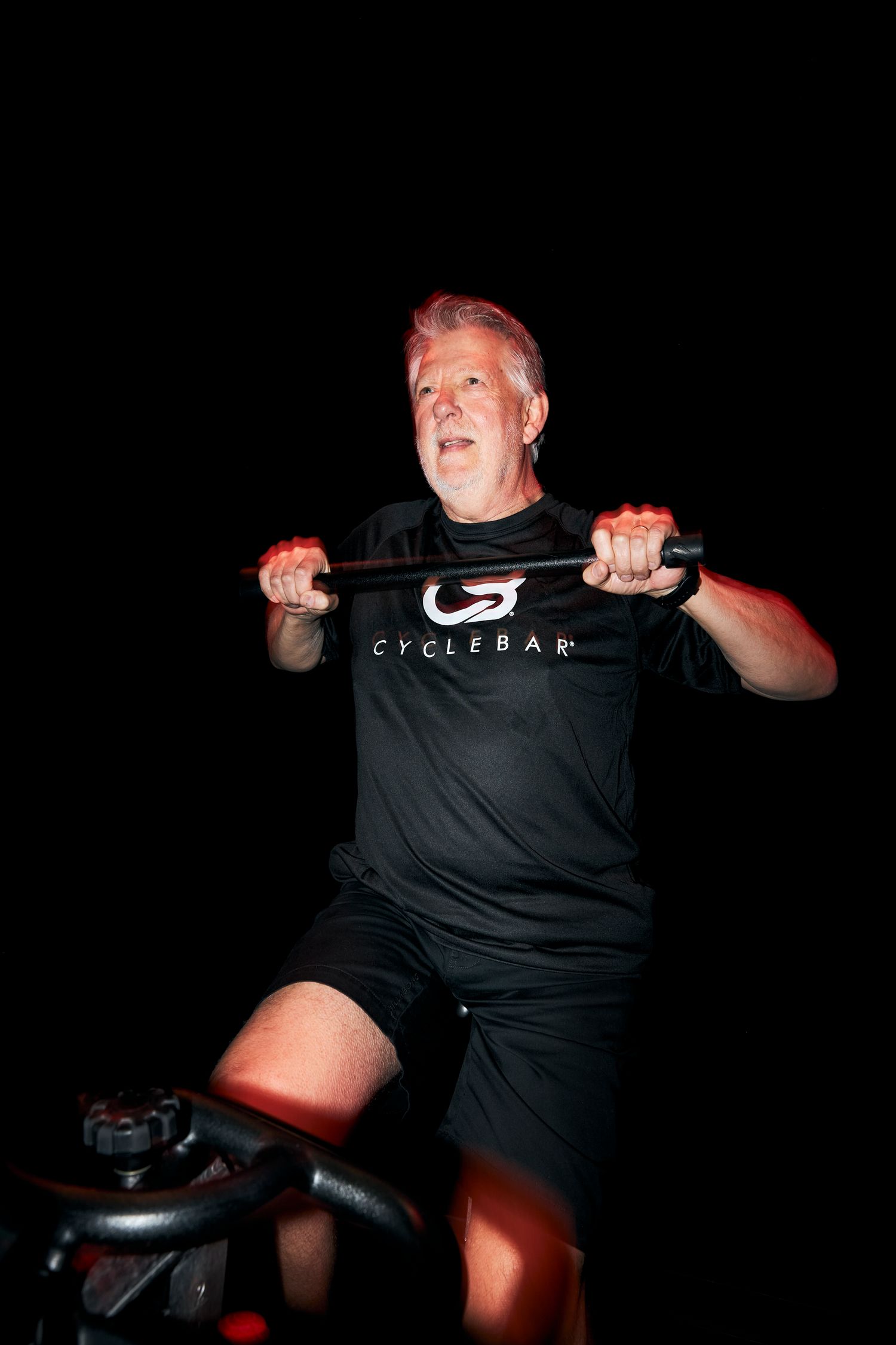 Older man riding a stationary bike at CycleBar indoor cycling studio, wearing a black CycleBar t-shirt.