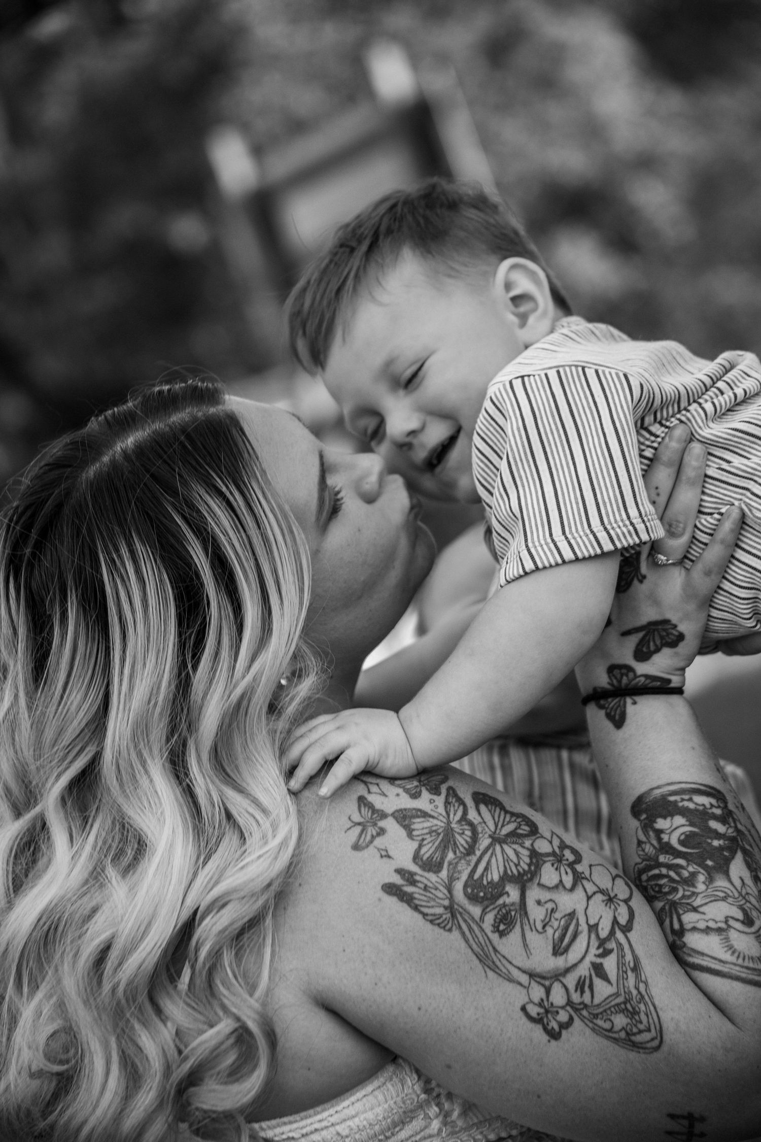 Black and white photo of a mother with tattoos lovingly holding her laughing baby boy close to her face.