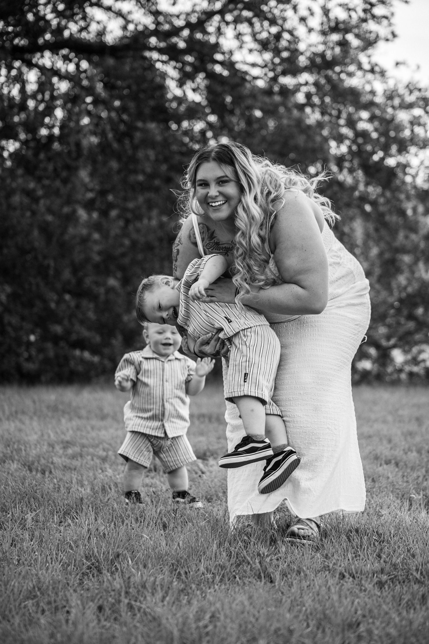 Black and white photo of a smiling woman holding two young boys in striped outfits on a grassy lawn.