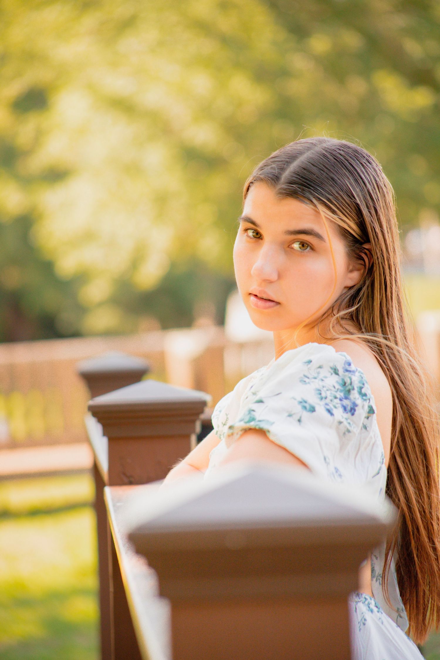 Young woman with long dark hair in floral dress leaning on wooden fence in sunny park setting.