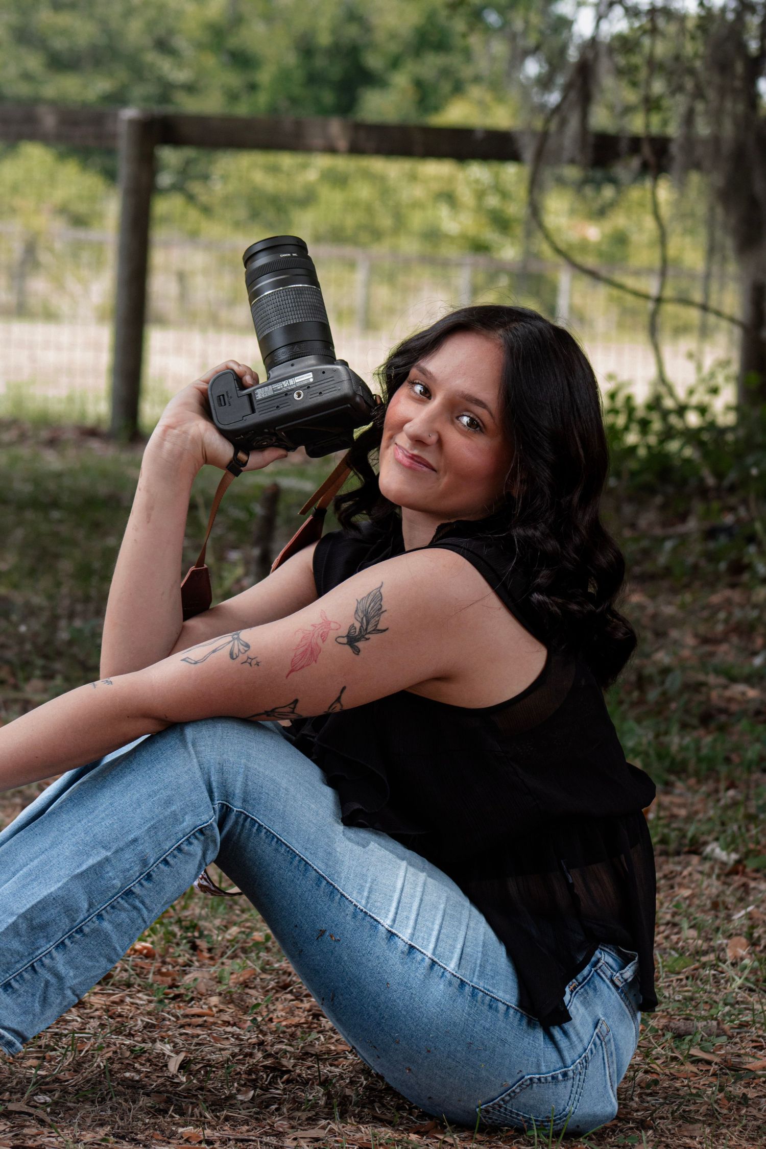 Young woman with tattoos sitting outdoors holding a DSLR camera, smiling, wearing jeans and black top near a fence.