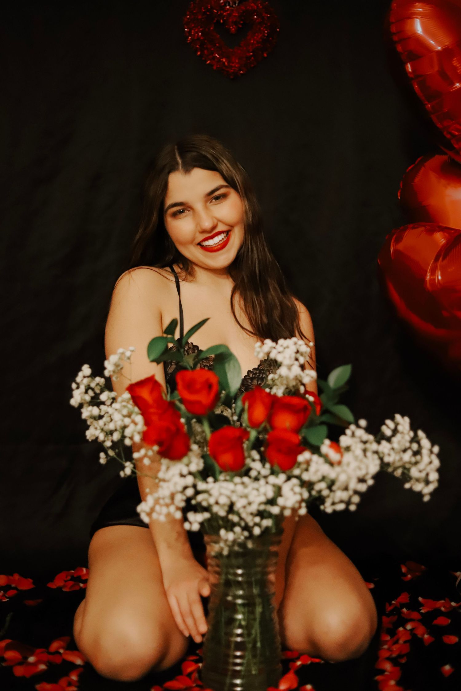 Smiling young woman with red lips kneels beside a vase of red roses with heart balloons in background.
