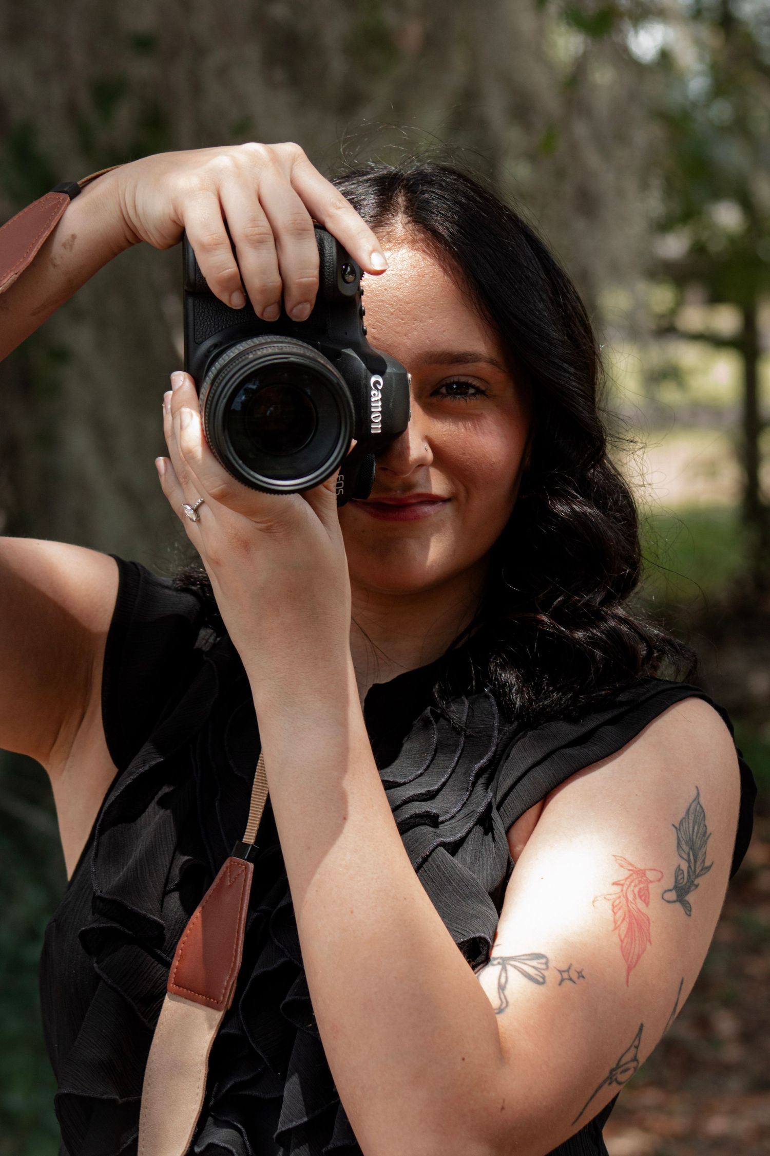 Woman with dark hair holding a Canon DSLR camera up to her eye, wearing a black dress with a floral tattoo on her arm.