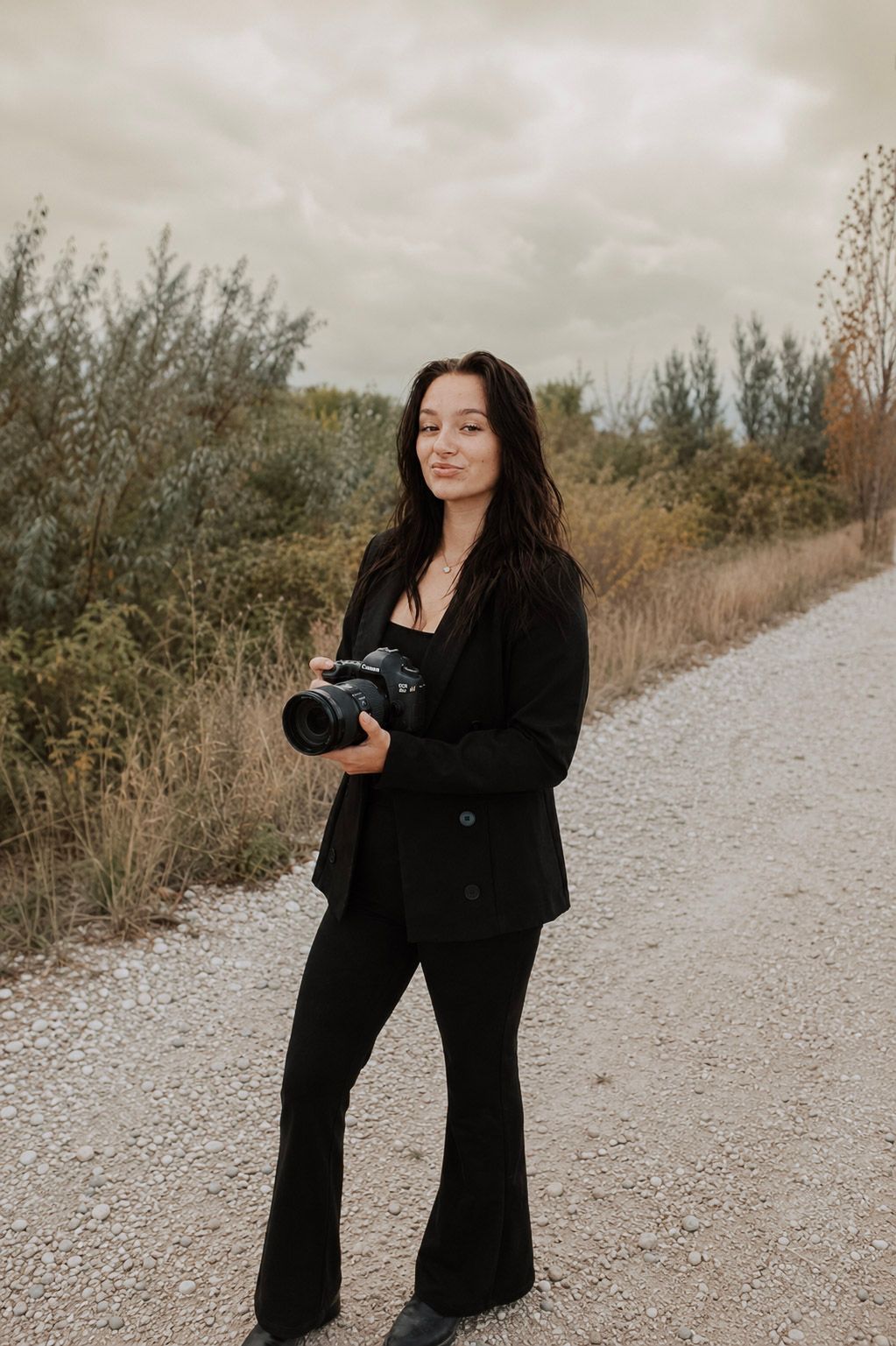 Young woman dressed in all black holds a camera while standing on a gravel path with autumn trees behind her.