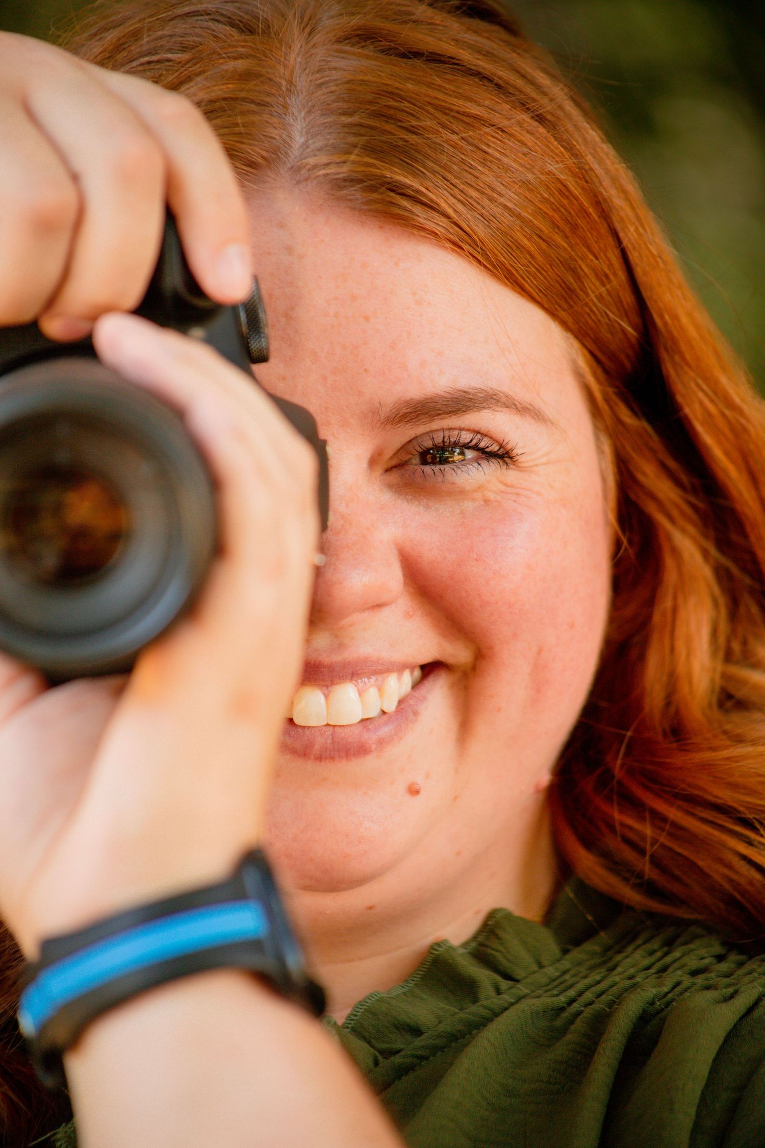Smiling redhead woman holding a DSLR camera up to her eye, wearing a green top and blue wristband outdoors.