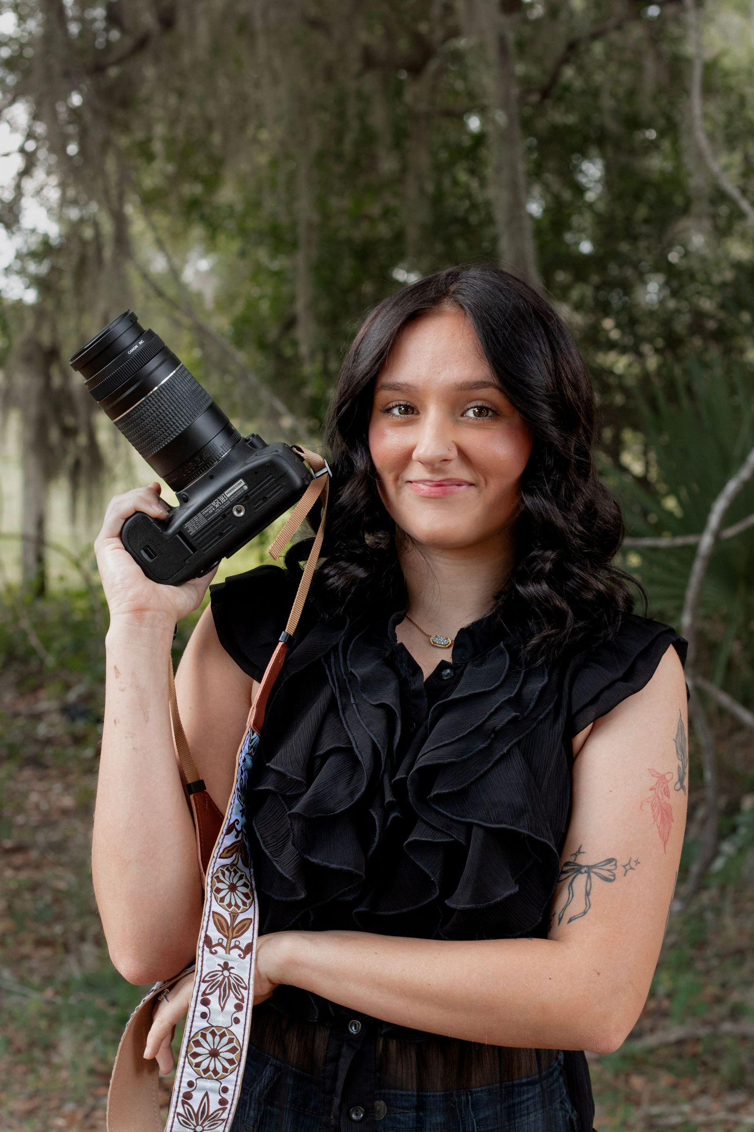 A smiling female photographer with dark wavy hair holds a DSLR camera outdoors, wearing a black ruffled top with a tattoo on her arm.