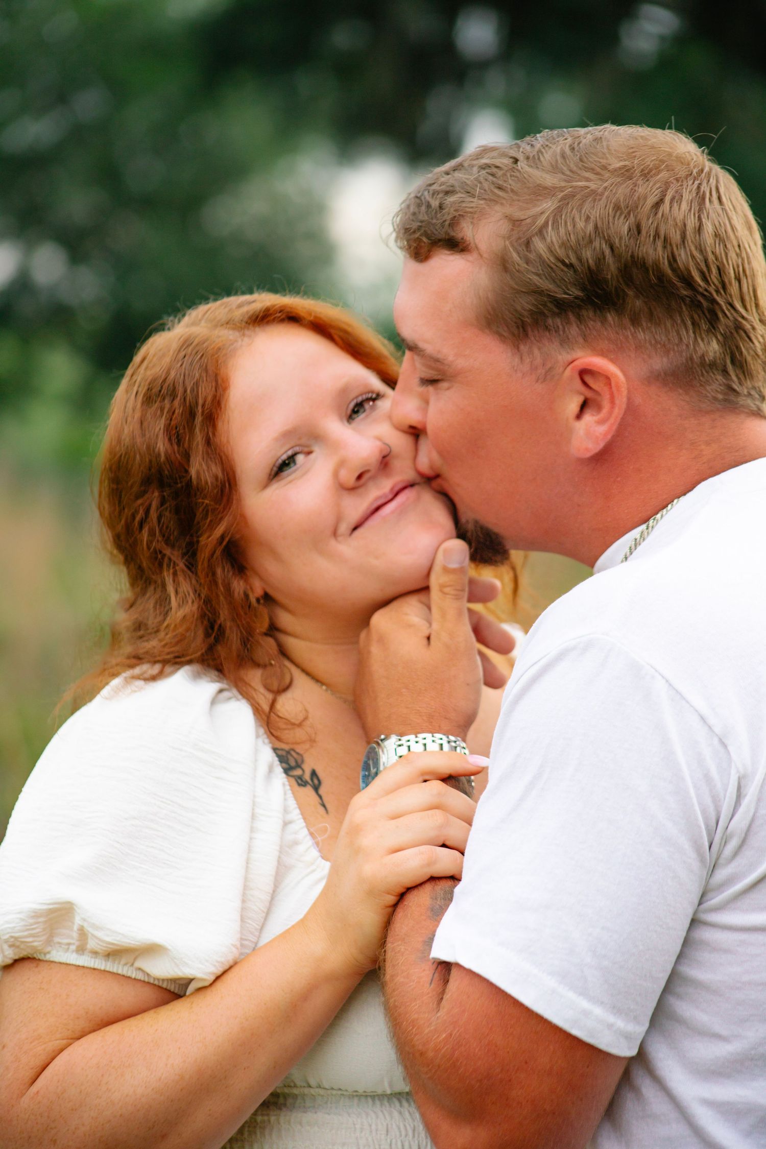 A man in a white t-shirt kisses a smiling redhead woman on the cheek outdoors.