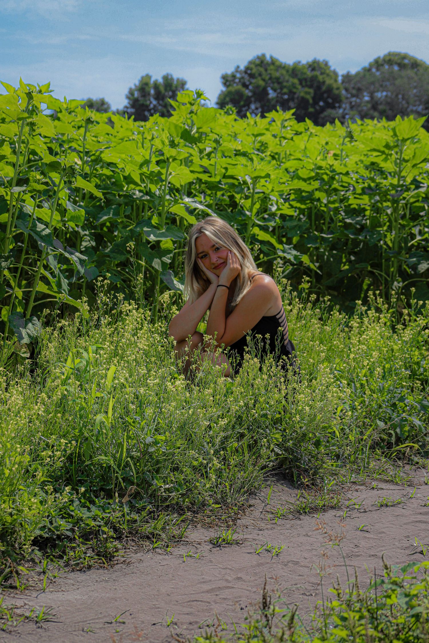 Blonde woman in black dress crouching among lush green plants and wildflowers in a sunny field.