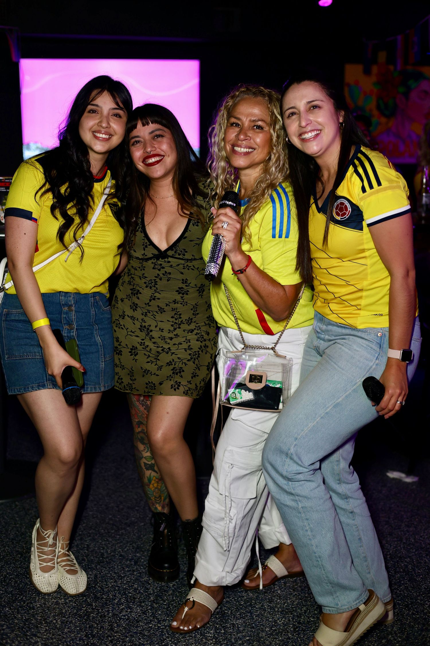Four women posing together at an event, three wearing yellow Colombia soccer jerseys, one holding a microphone.