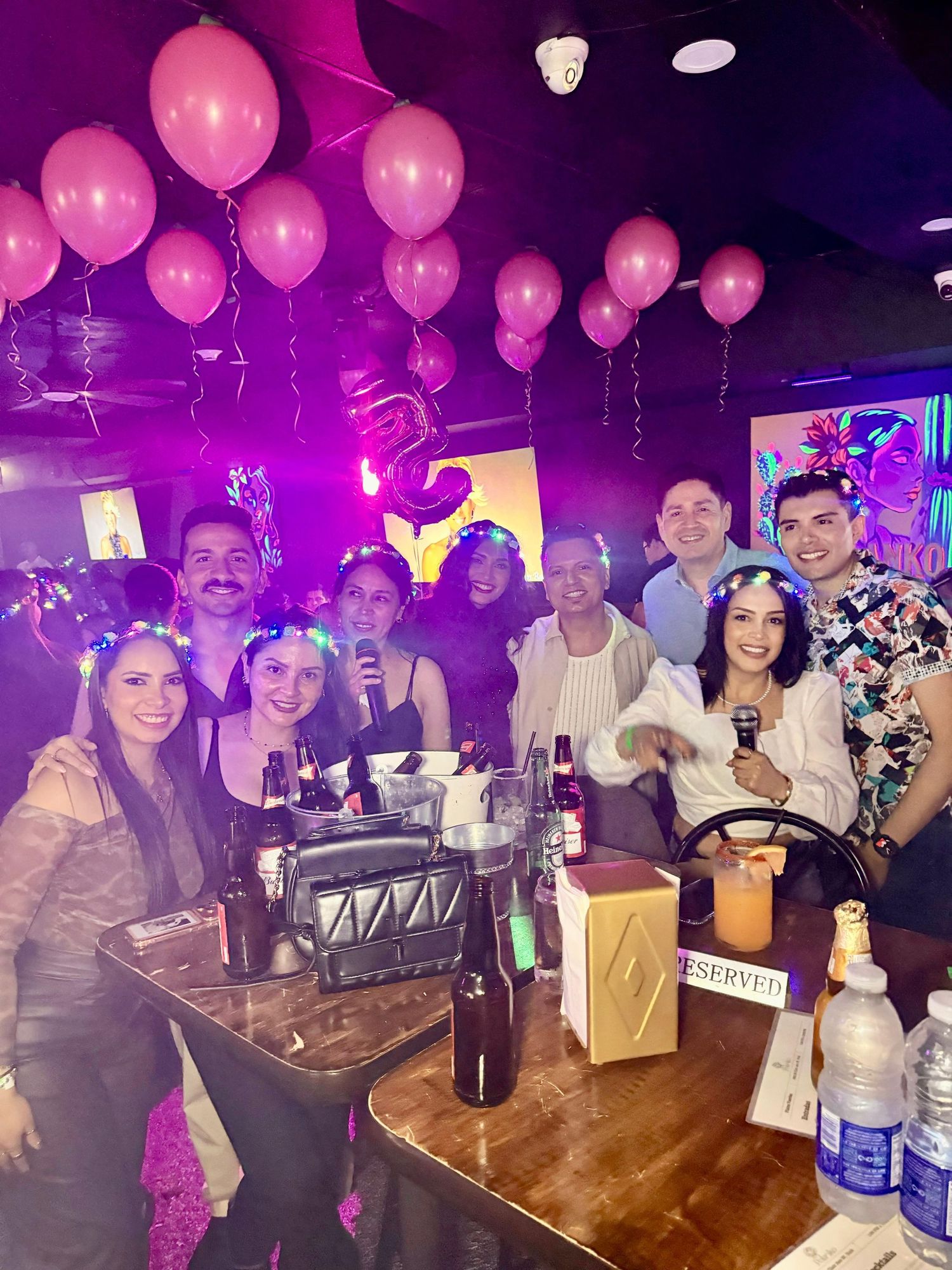Friends enjoying drinks and pink balloons at a nightclub with purple lighting.