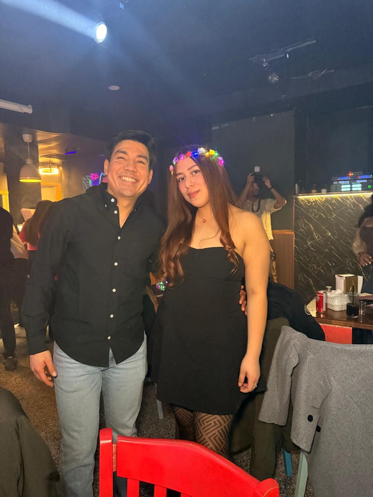 Two people pose together at a nightclub, one wearing a black shirt and the other in a black dress with a colorful flower crown.