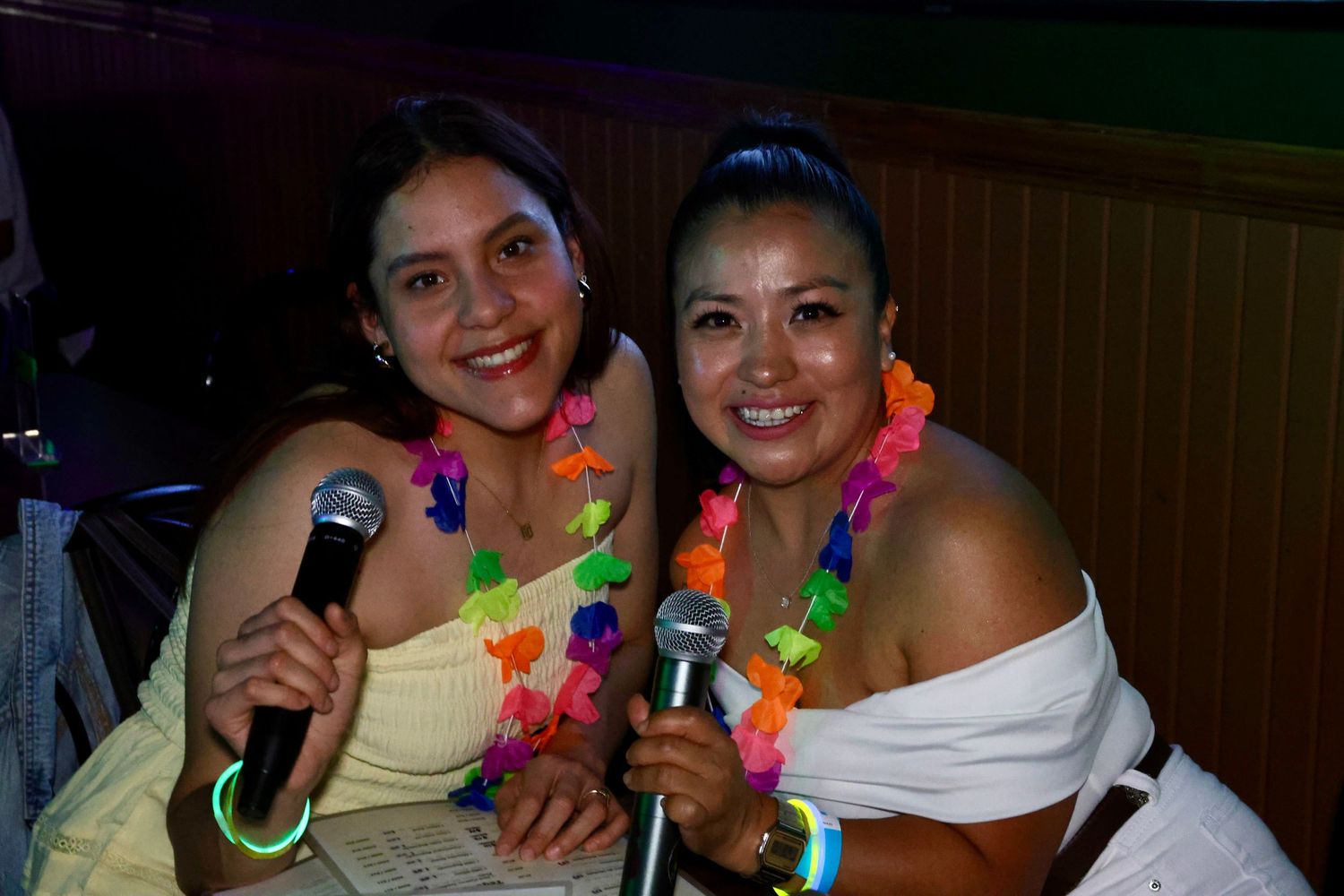 Two women in white outfits wearing colorful lei necklaces smile while holding microphones at a karaoke event.