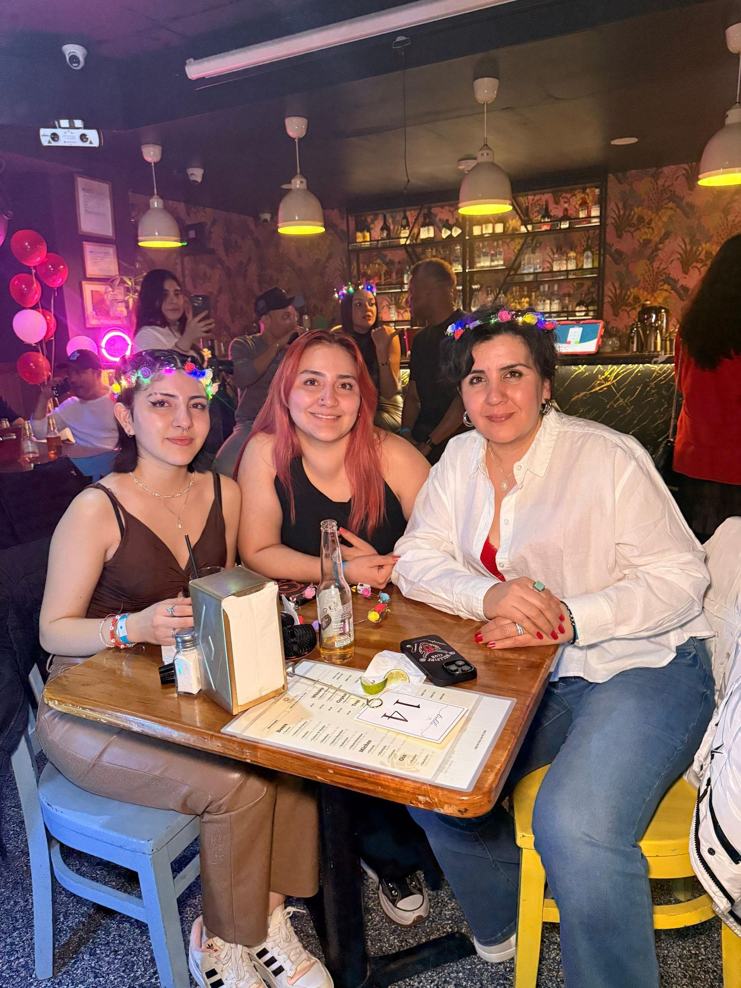 Three friends share drinks and conversation at a cozy bar table with pendant lighting overhead.