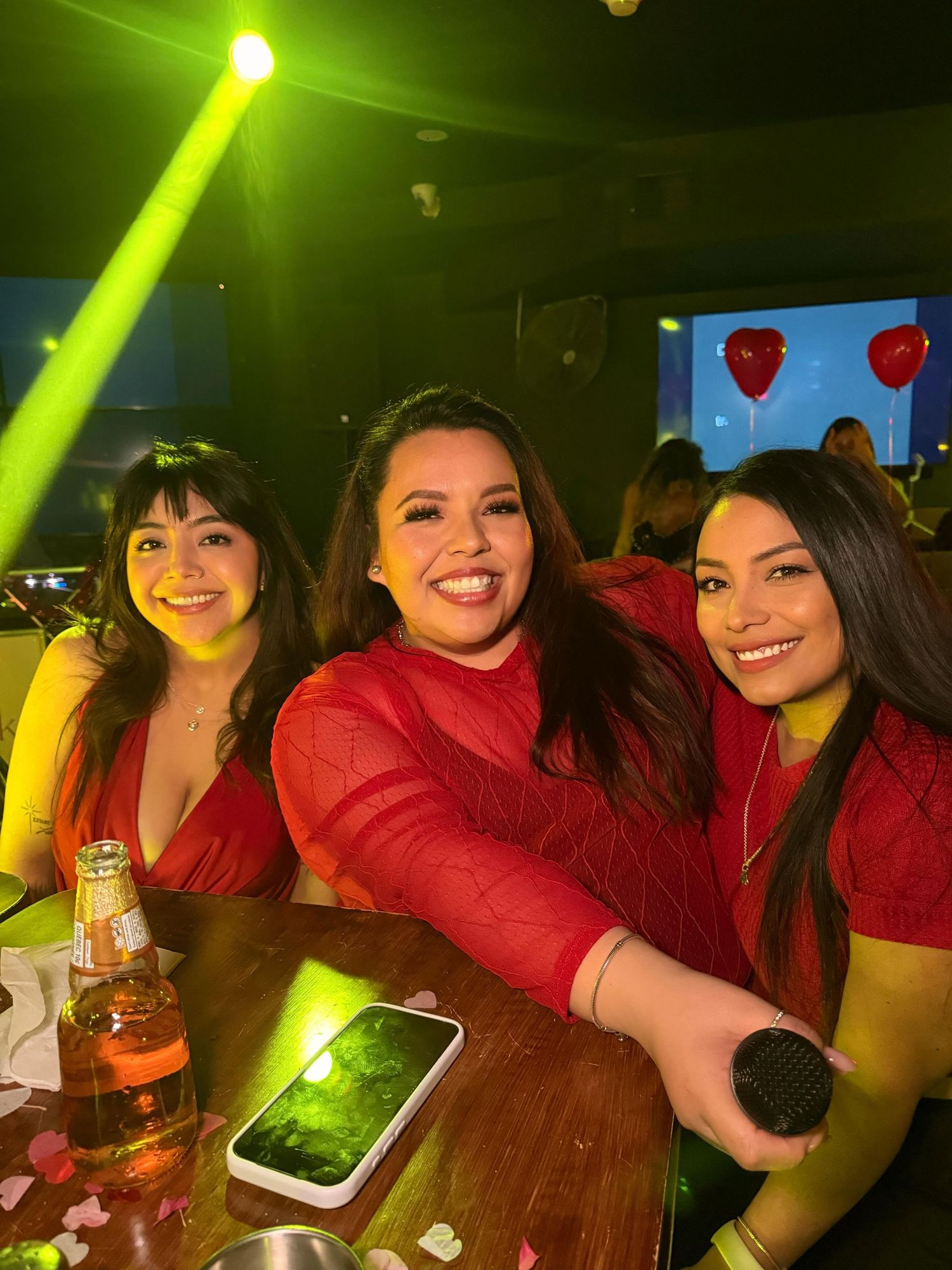 Three friends in red outfits enjoying drinks at a bar with green lighting.