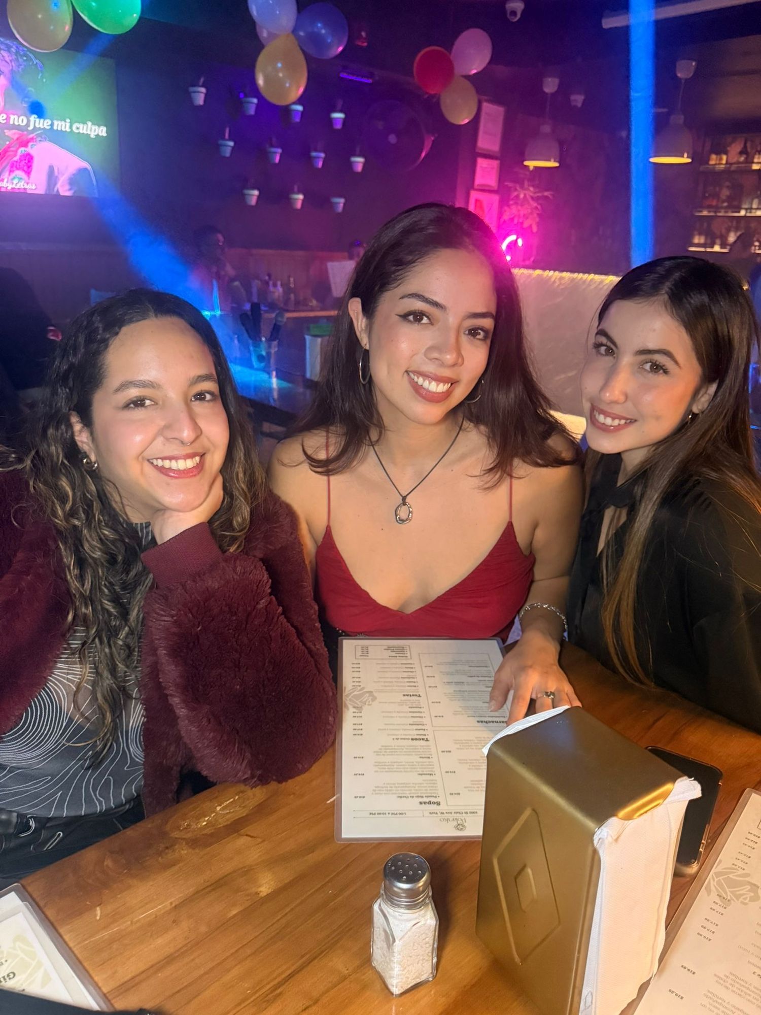 Three smiling women pose together at a restaurant booth with colorful balloons and neon lights in the background.