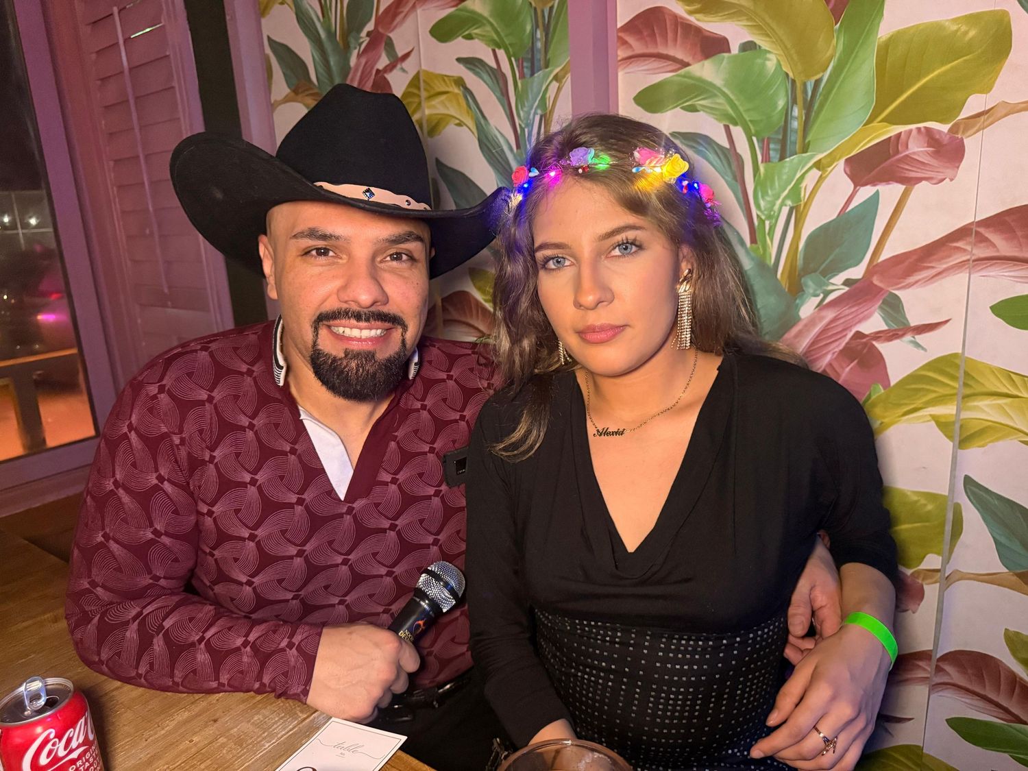 A couple poses at a restaurant with tropical leaf wallpaper backdrop and festive accessories.